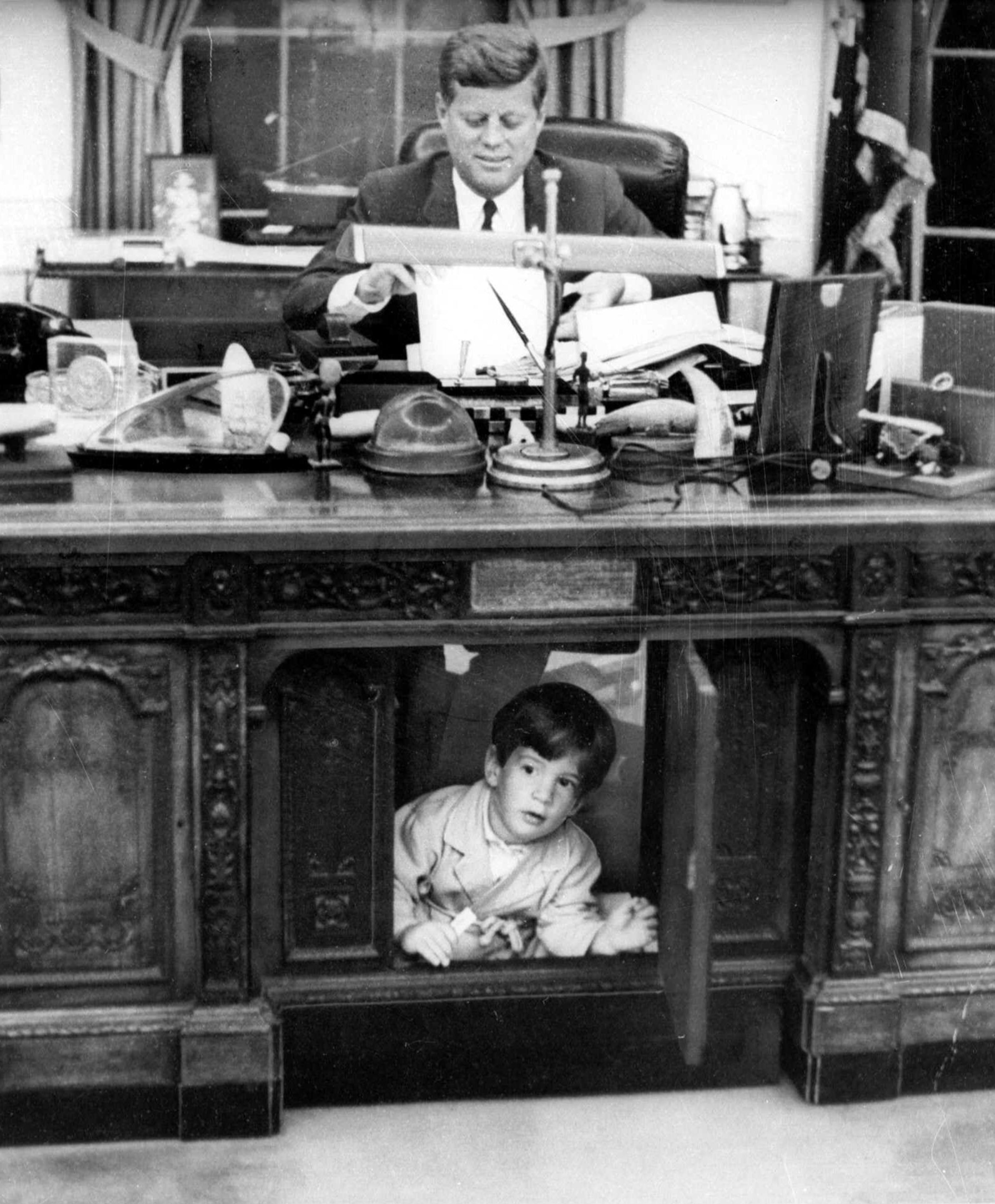 President Kennedy, working late at his White House office, wears a slight smile on his face, indicating perhaps he is not completely unaware that his son, John Jr., is exploring under his desk in the Oval Office in the White House in 1963. John Jr. called the spot under the desk 'my house' and was peeking from behind the 'secret door.' A small plane carrying John F. Kennedy Jr. to a wedding in Hyannisport, Mass., was reported missing early Saturday, July 17, 1999, and a search was under way off the coast of New York's Long Island, Coast Guard officials said. (AP Photo/Look Magazine, Stanley Tretick, File)