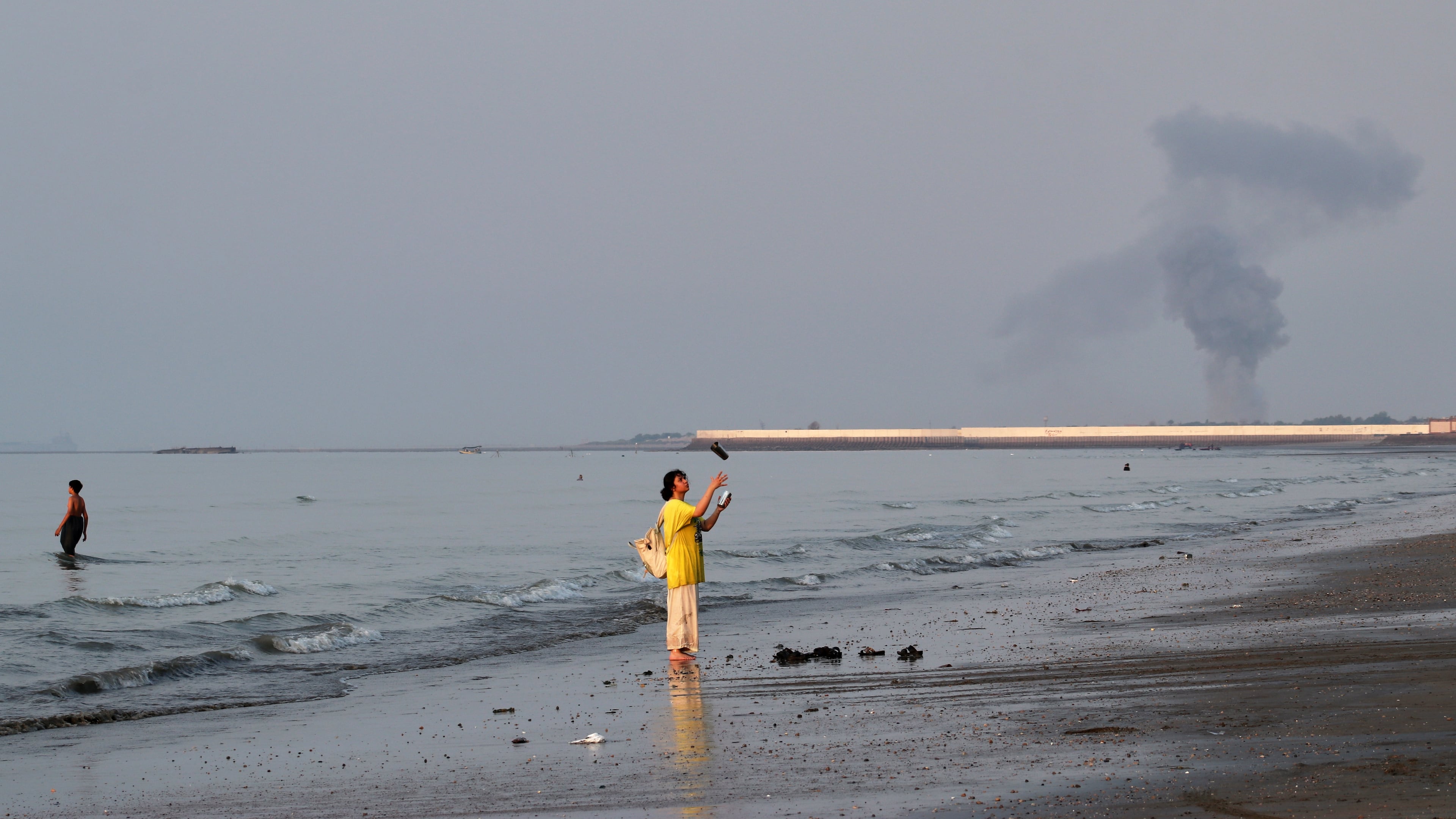 A woman stands at the water's edge along the Strait of Hormuz as a plume of smoke rises in the background following an explosion, off Bandar Abbas, Iran, Monday, July 13, 2026. (Razieh Poudat/ISNA via AP)