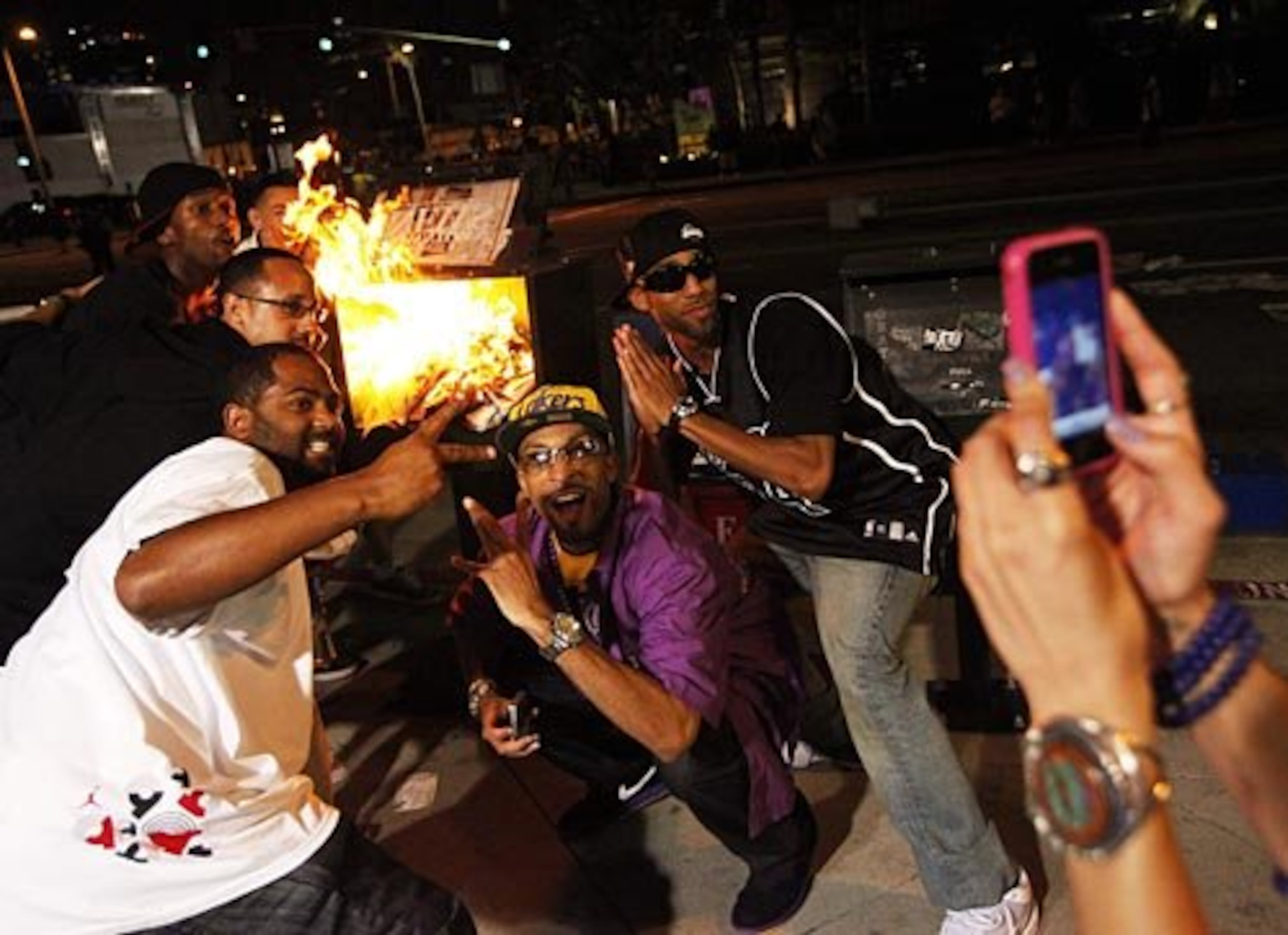 Lakers fans pose by a burning newspaper stand as another person takes a photo using a cellphone camera.