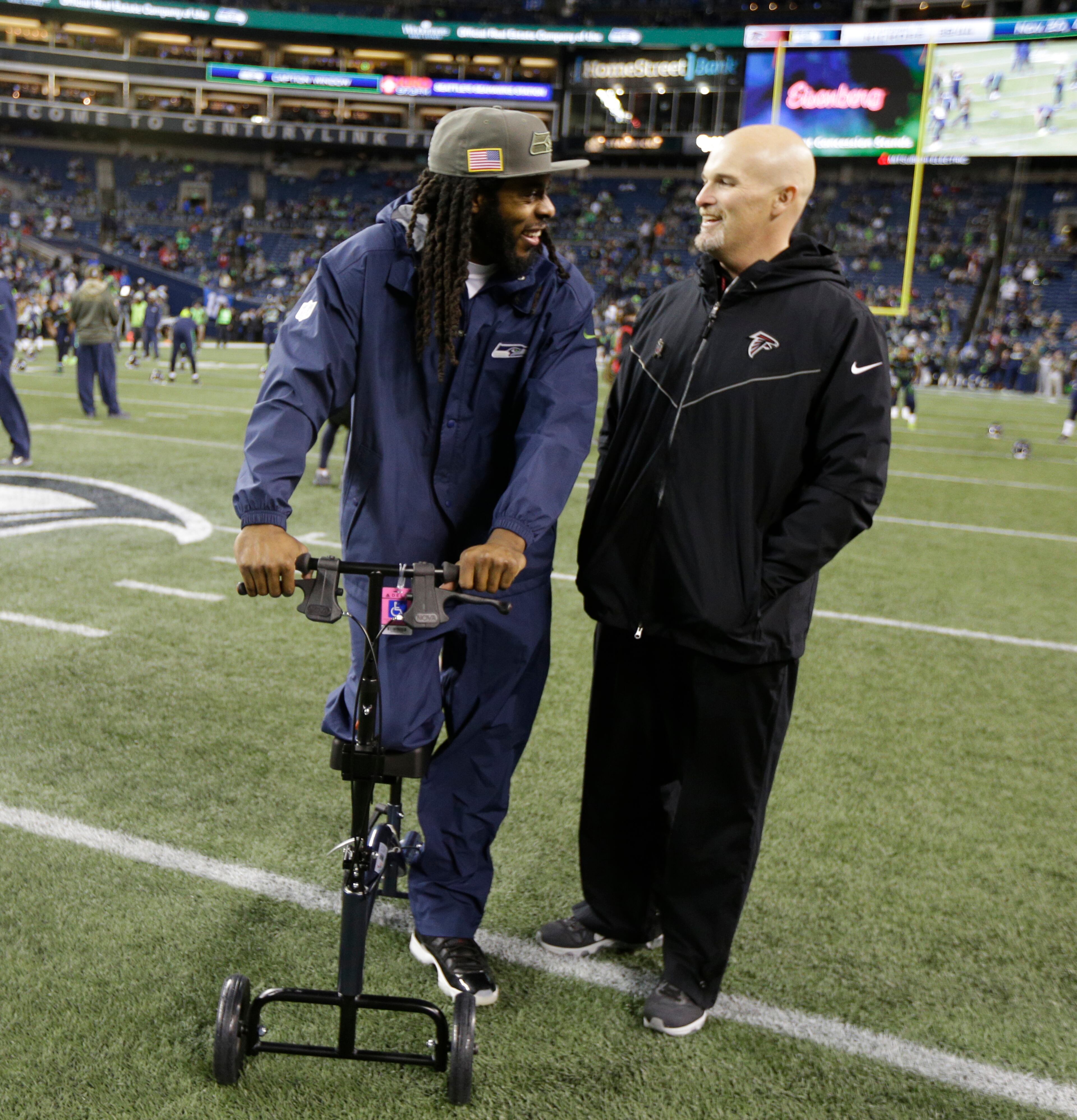 Seattle Seahawks' Richard Sherman, left, leans on a cart protecting his injured leg as he greets Atlanta Falcons head coach Dan Quinn before an NFL football game, Monday, Nov. 20, 2017, in Seattle. (AP Photo/Ted S. Warren)
