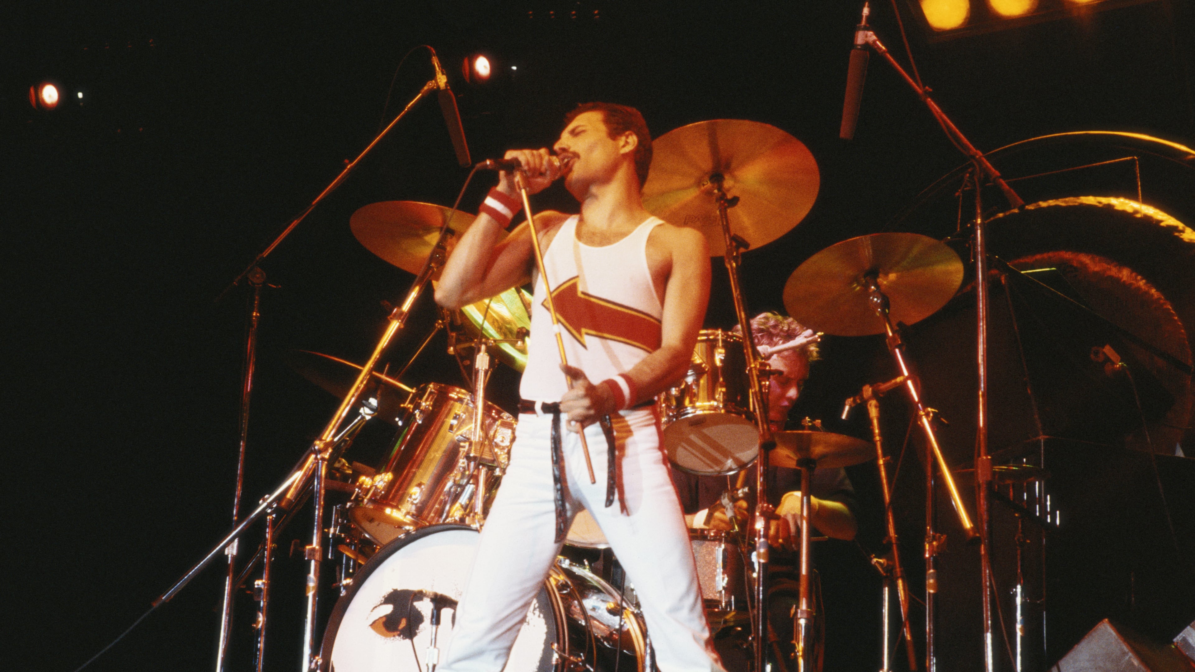 Freddie Mercury (1946-1991), singer with Queen, standing in front of a drumkit as he sings into a microphone on stage during a live concert performance by the band at the National Bowl in Milton Keynes, England, United Kingdom, on 5 June 1982. The band's anthem, "We Will Rock You" remains a staple at sporting events.