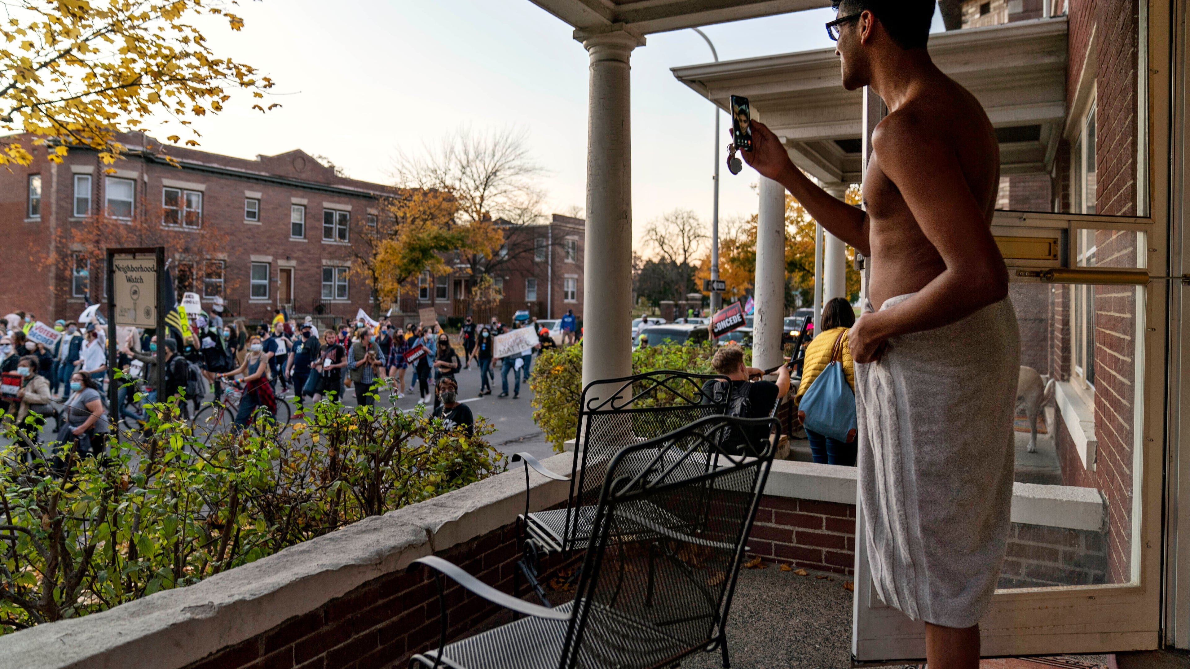 FILE - Karthik Mantha stands on his front porch wearing a towel, just as he was about to step into a shower, to watch a rally pass by celebrating the presidential election results in Detroit, Nov. 7, 2020. (AP Photo/David Goldman, File)