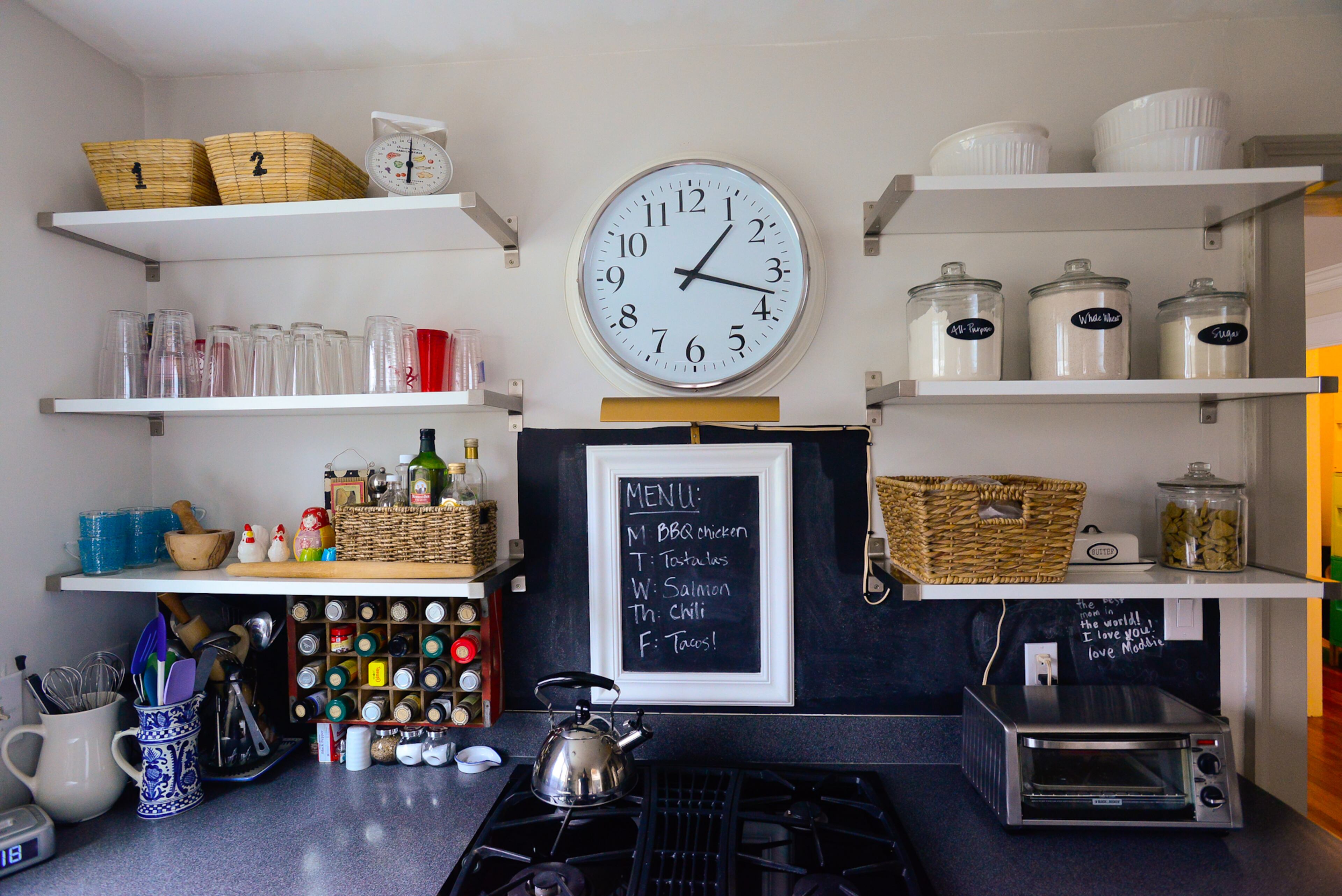 The homeowners replaced the upper cabinets with IKEA shelves that are easier to access. The backsplash was painted with Rust-Oleum chalkboard paint, the clock is from IKEA and the spice rack to the left of the stove was made from an old Coca-Cola crate.