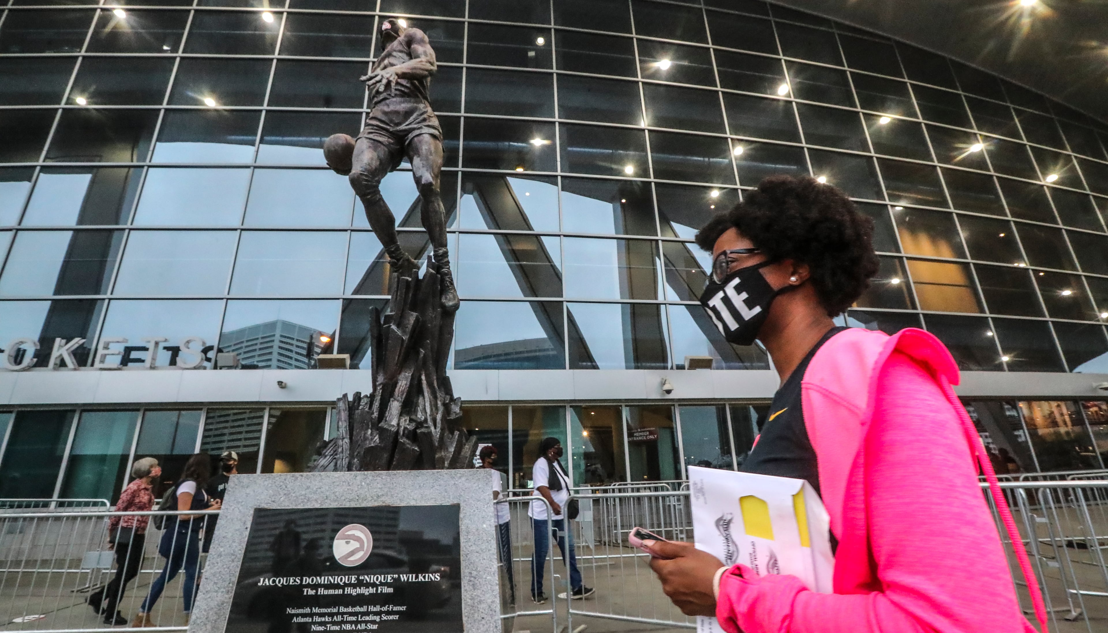 October 12, 2020 Atlanta: Ashley Nealy makes her way past the statue of Dominique Wilkins as she waited to vote on Monday, Oct. 12, 2020 at State Farm Arena in downtown Atlanta. Eager Georgia voters swarmed to polling places Monday morning, waiting in lines created by high turnout and technical problems at the start of three weeks of early voting before Election Day. A glitch with voter check-in computers held up voters at Georgia’s largest early voting site at State Farm Arena. Lines stopped after voters received an “invalid card” error when inserting green voter access cards into touchscreens. Poll workers had to reboot the arena’s 60 voter check-in tablets and re-import voter information, said Fulton Elections Director Richard Barron. “We apologize to all the voters,” Barron said. (John Spink / John.Spink@ajc.com)