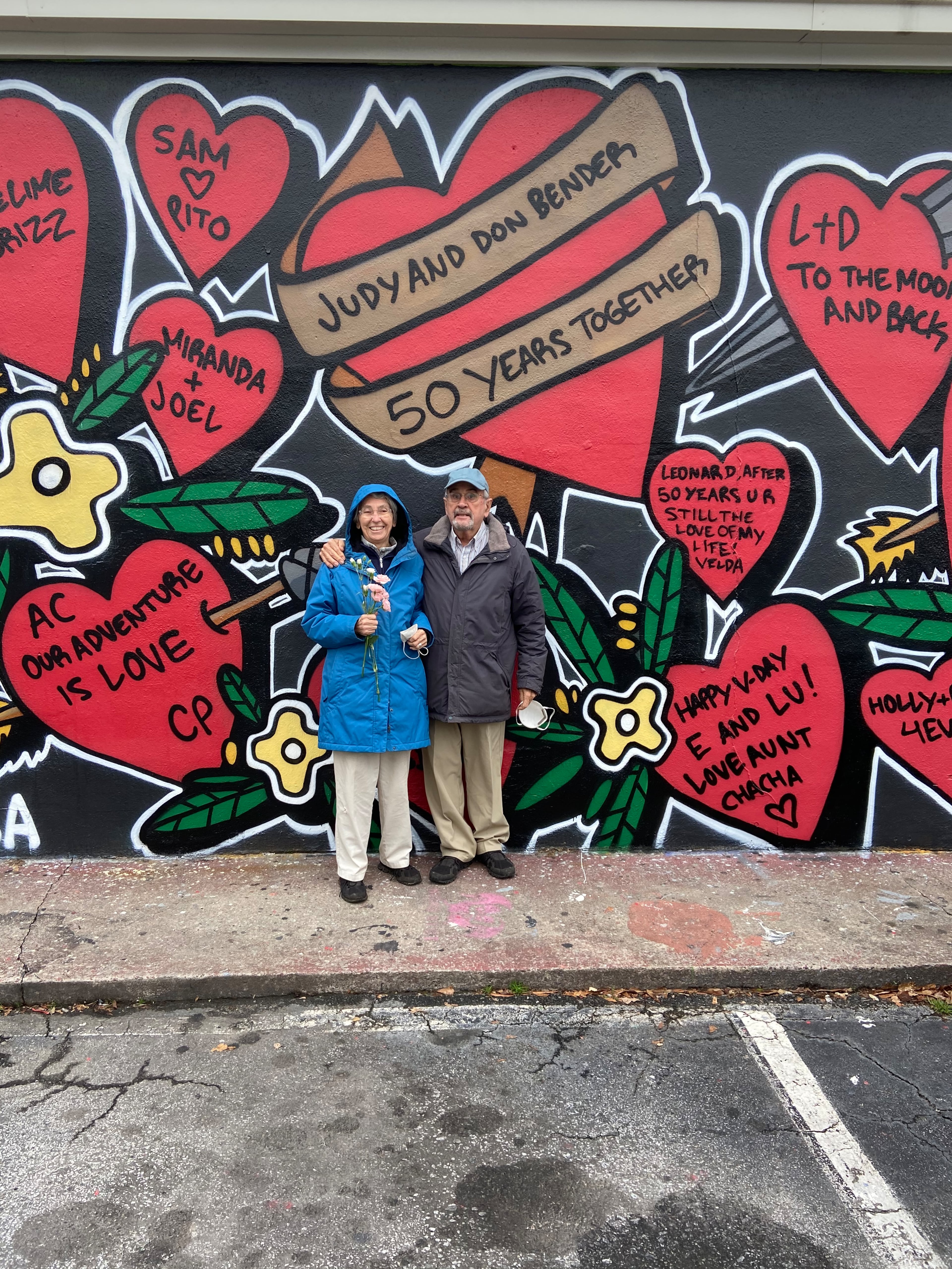 Judy Bender (left) holds flowers given to her by her husband Don Bender (right) while posing underneath a giant heart Don sponsored as part of the annual Little Five Points Valentine's Day mural. The couple was celebrating 50 years of marriage. The mural serves as a fundraiser for the Little 5 Points Cultural District and helps Atlantans surprise their loved ones with a public display of affection. (Kelly Stocks photo)