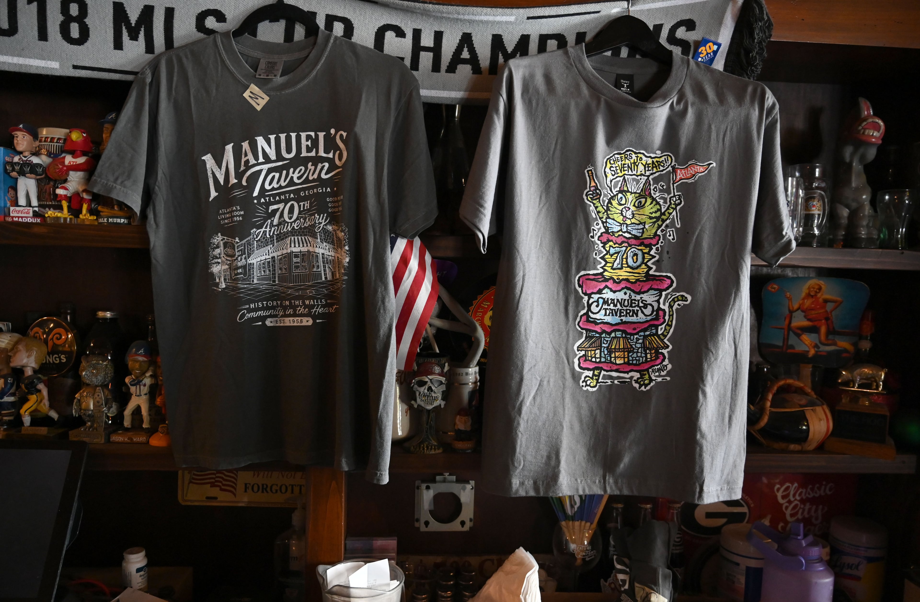 Two 70th anniversary T-shirts, including a classic design and another created by renowned intown artist R. Land, are offered for sale at Manuel’s Tavern on Thursday, Aug. 6, 2026, in Atlanta. (Hyosub Shin/AJC)