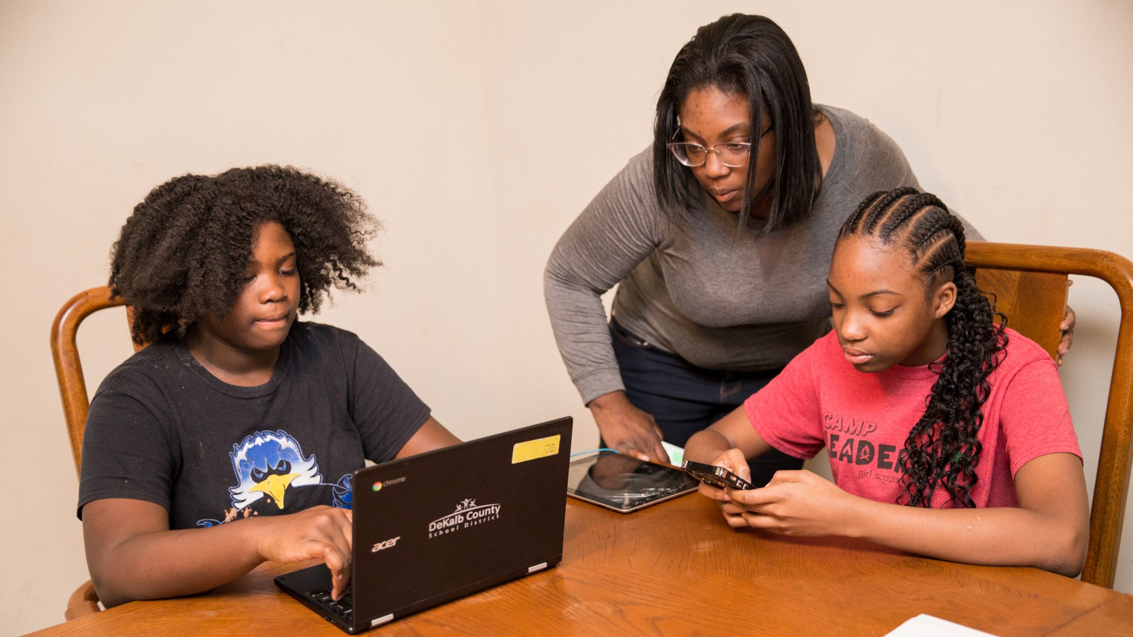 Online learning at the Hill house in Lithonia starts just after 5 pm when their mom gets home from her job at Lowe’s. Raina Hill, 12, left, a 6th grader at Champion Middle School logs in with help from her mother, LaTonya Hill, standing, while Rihanna Hill, 13, in red, an 8th grader at Champion Middle School uses a tablet to do school work. (Jenni Girtman for Atlanta Journal Constitution)