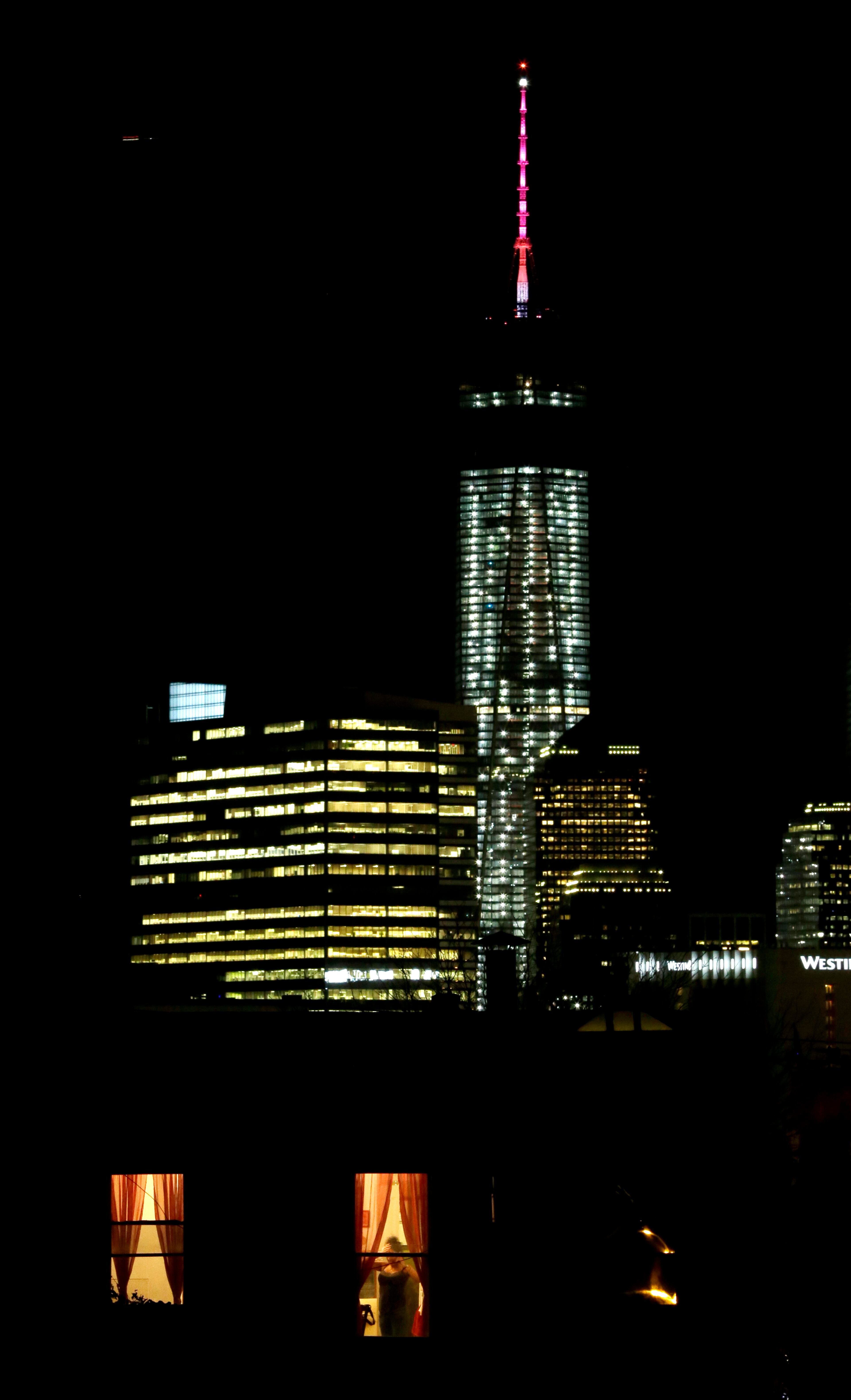 A woman talking on a phone is seen through a window of a home in The Heights neighborhood of Jersey City, N.J., with One World Trade Center as a backdrop, Friday, Nov. 8, 2013. The Port Authority of New York and New Jersey, who tested the lights on Friday, said the beacon is packed with nearly 300 modules and their glow can be seen for up to 50 miles. The beacon and spire together stand 408 feet tall and bring the building, formerly called the Freedom Tower, to its symbolic height of 1,776 feet. The Durst Organization operates the spire, which will serve broadcasters. (AP Photo/Julio Cortez)