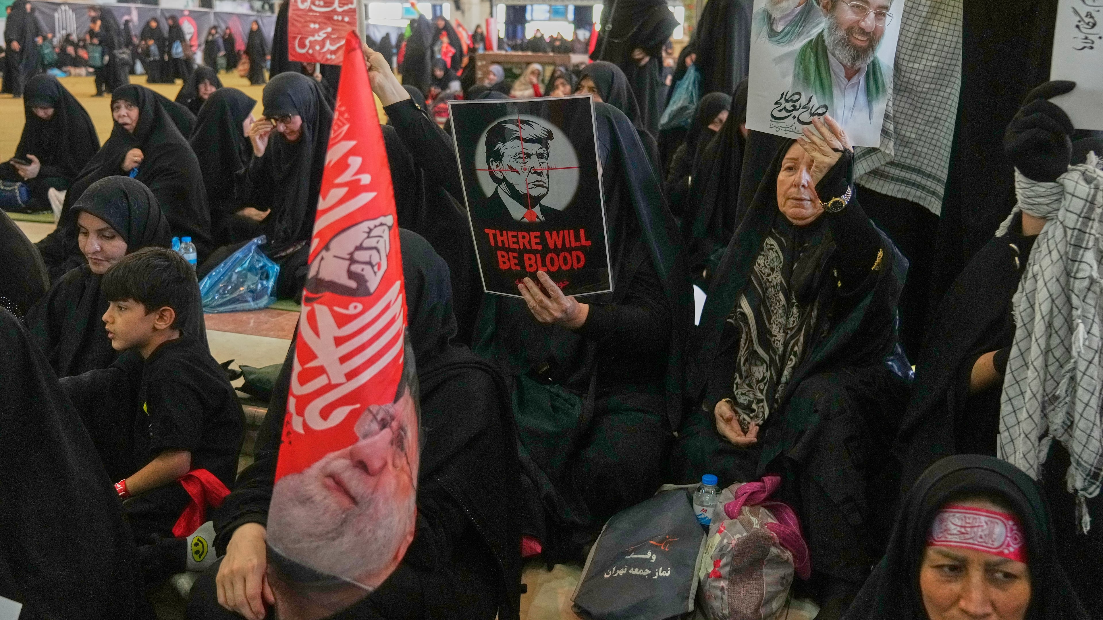 A woman holds a poster depicting U.S. President Donald Trump in the crosshairs of a rifle scope with the English words "There Will Be Blood," while another woman holds a portrait of the late Supreme Leader Ali Khamenei and his son, current Supreme Leader Mojtaba Khamenei, as mourners gather to commemorate the late leader at the Imam Khomeini Mosalla Grand Mosque in Tehran, Iran, Tuesday, July 14, 2026. (AP Photo/Vahid Salemi)