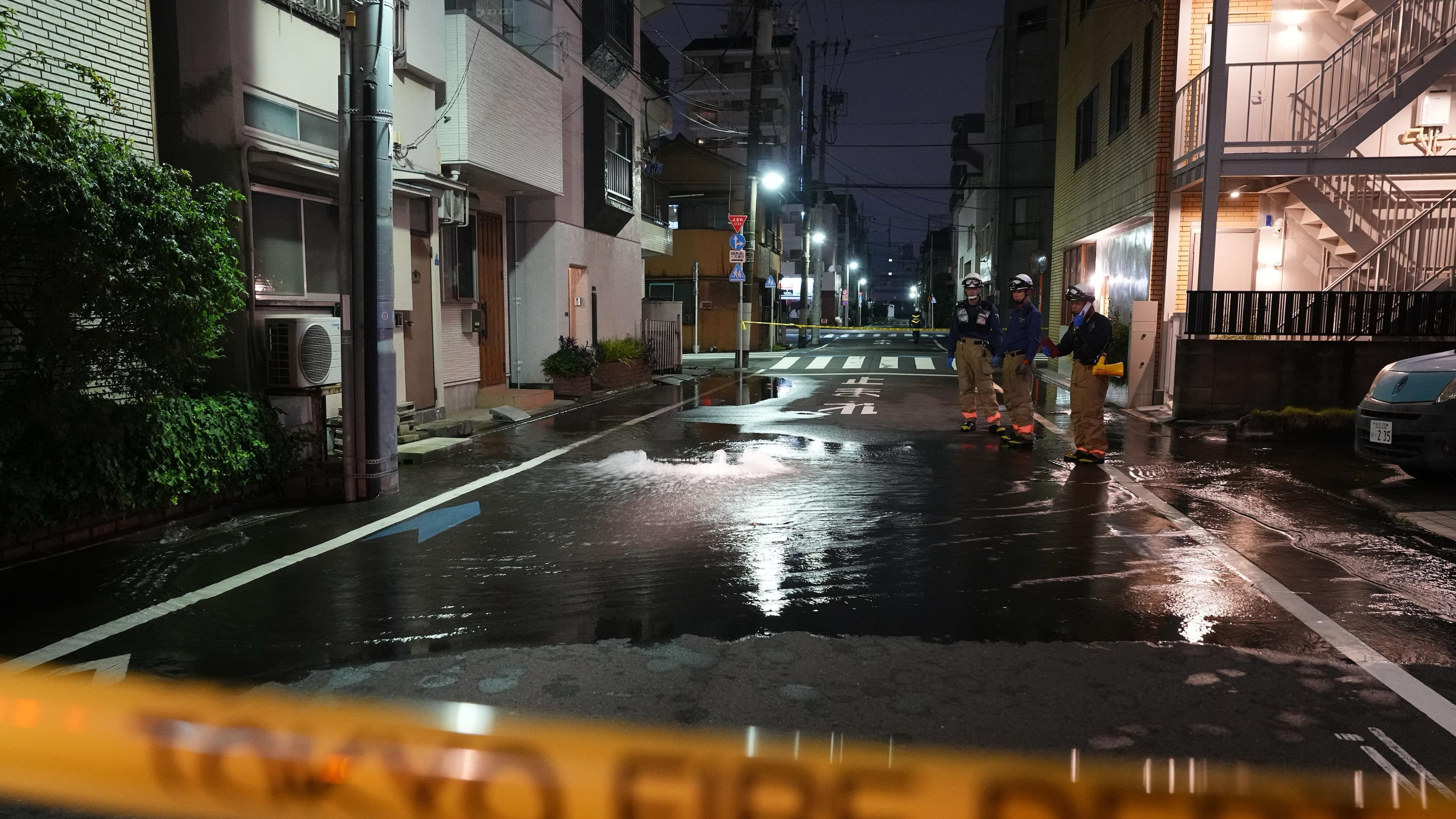 First responders from the Tokyo Fire Department stand on a street partially flooded in the early hours of Sunday, Aug. 23, 2026, following the rupture of a water pipe after an earthquake that struck the area and neighboring prefectures. (Kazuki Kozaki/Kyodo News via AP)