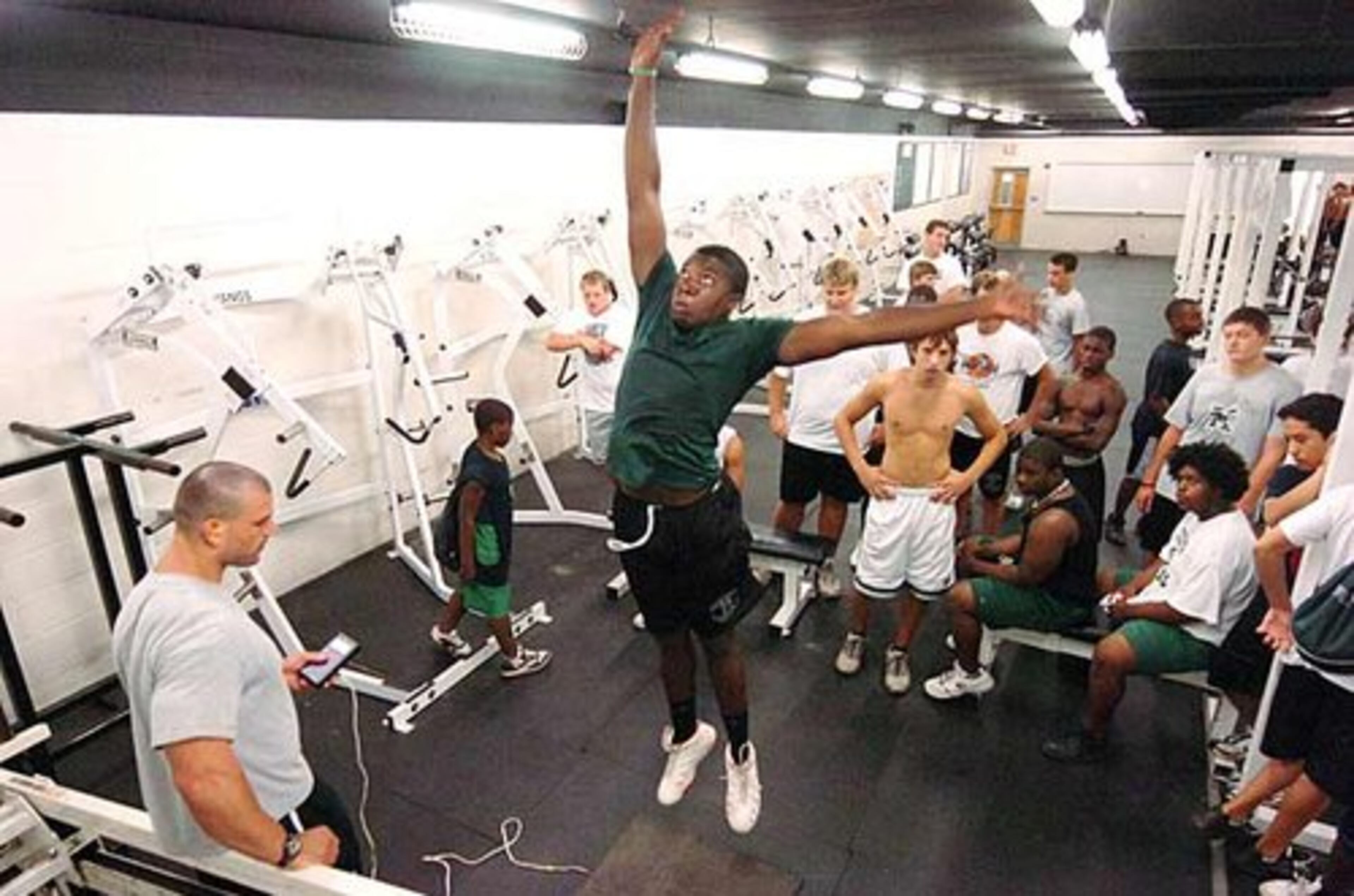 Mustangs strength coach Zach Reed (left) watches as senior DE Bryan Hill (in green shirt) does a big jump in the weight room during skills testing assessments.