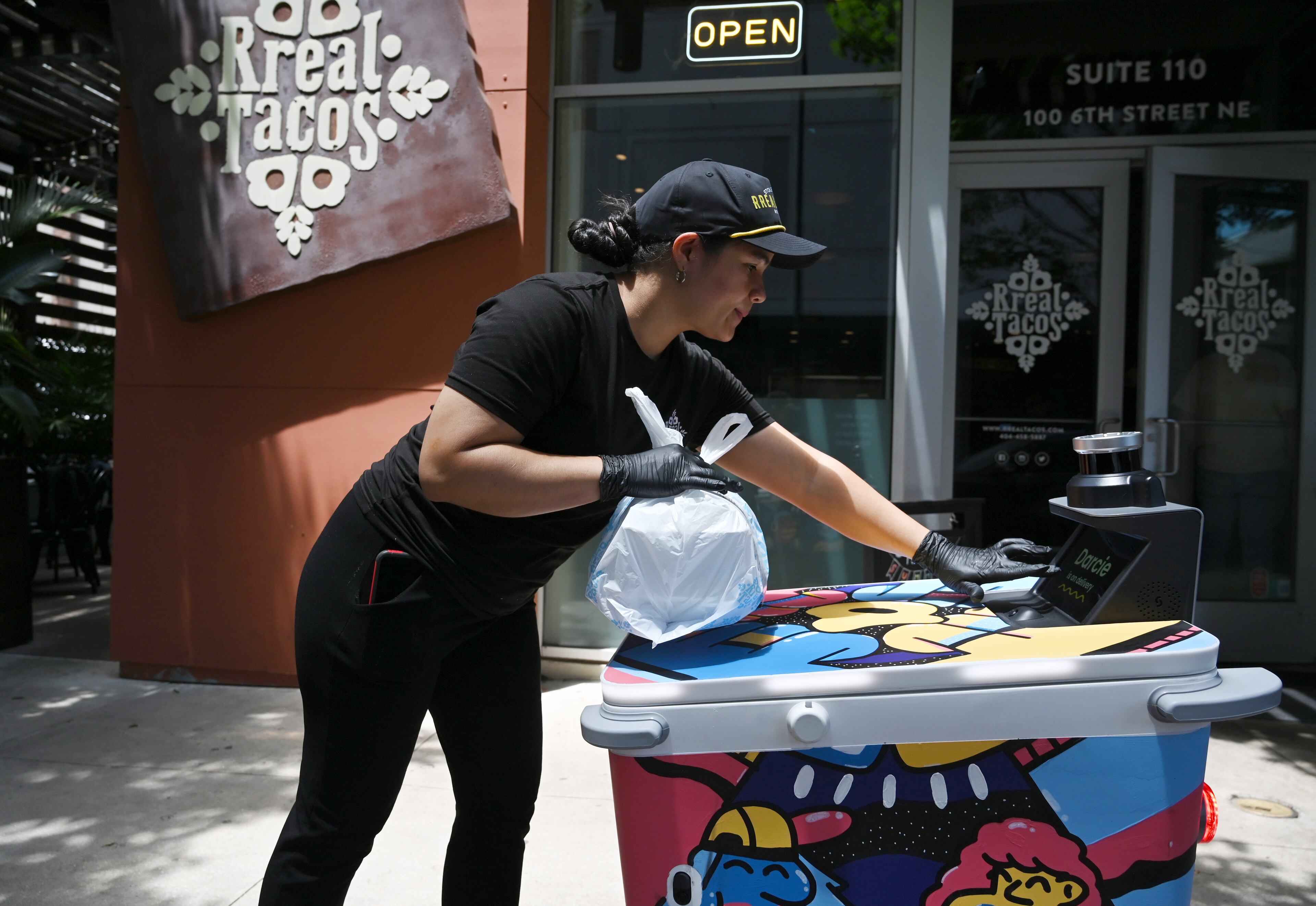 Patricia Sulbaran, staff at Rreal Tacos, places a food order inside delivery robot Darcie made by Serve Robotics. The robot was painted by Atlanta muralist George F. Baker III. (Hyosub Shin/AJC)