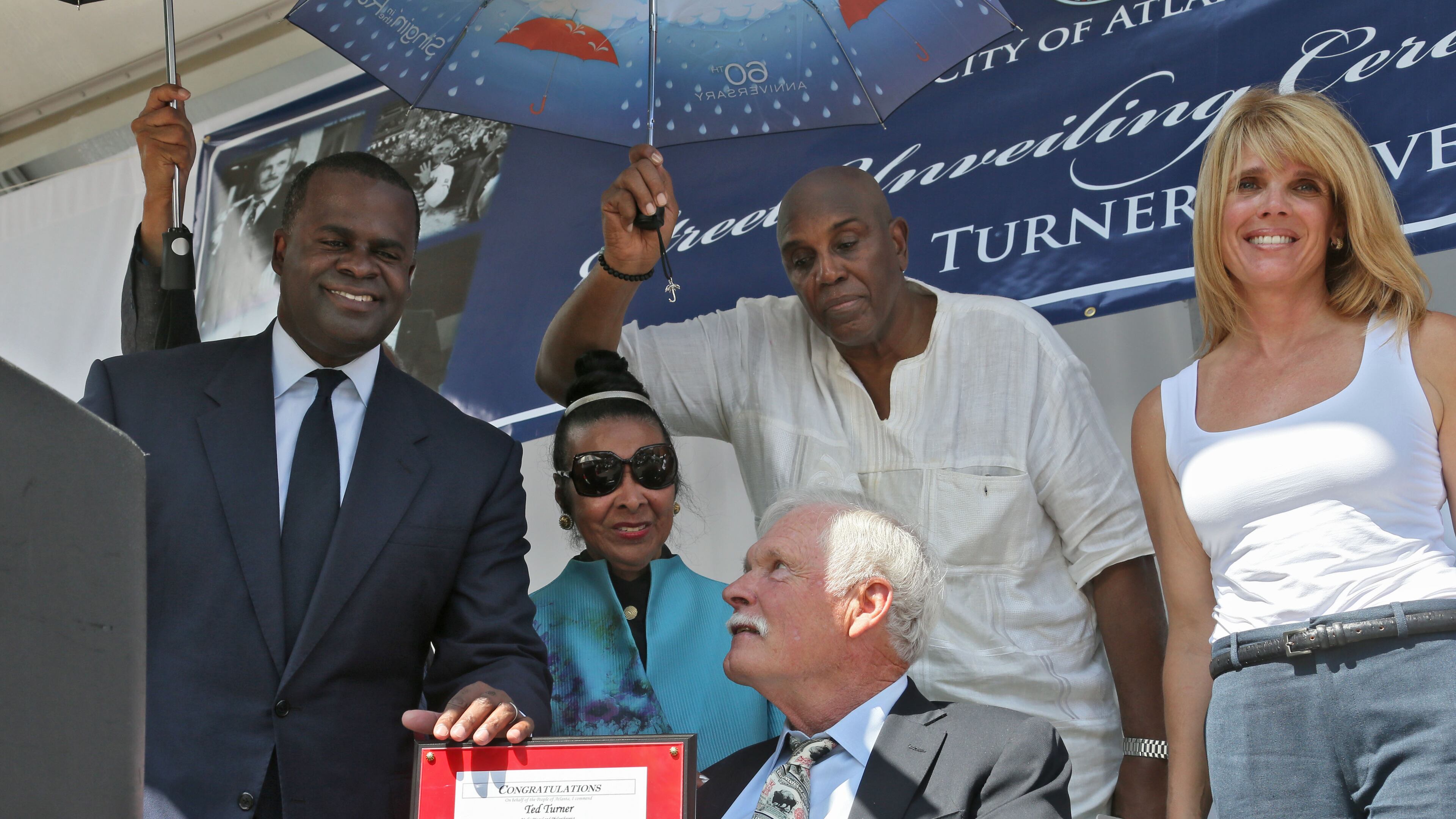 Seated under a canopy of umbrellas to shield the hot sun, Ted Turner receives a commendation from Mayor Kasim Reed (left). At right is his daughter, Laura Turner Seydel, Mayor Kasim Reed unveiled Ted Turner Drive on Tuesday morning. The renamed portion of Spring Street between Whitehall Street and West Peachtree Street is near the area where Turner established some of his most prominent business endeavors, including Turner Broadcasting System (TBS) and the CNN Headquarters. BOB ANDRES / BANDRES@AJC.COM