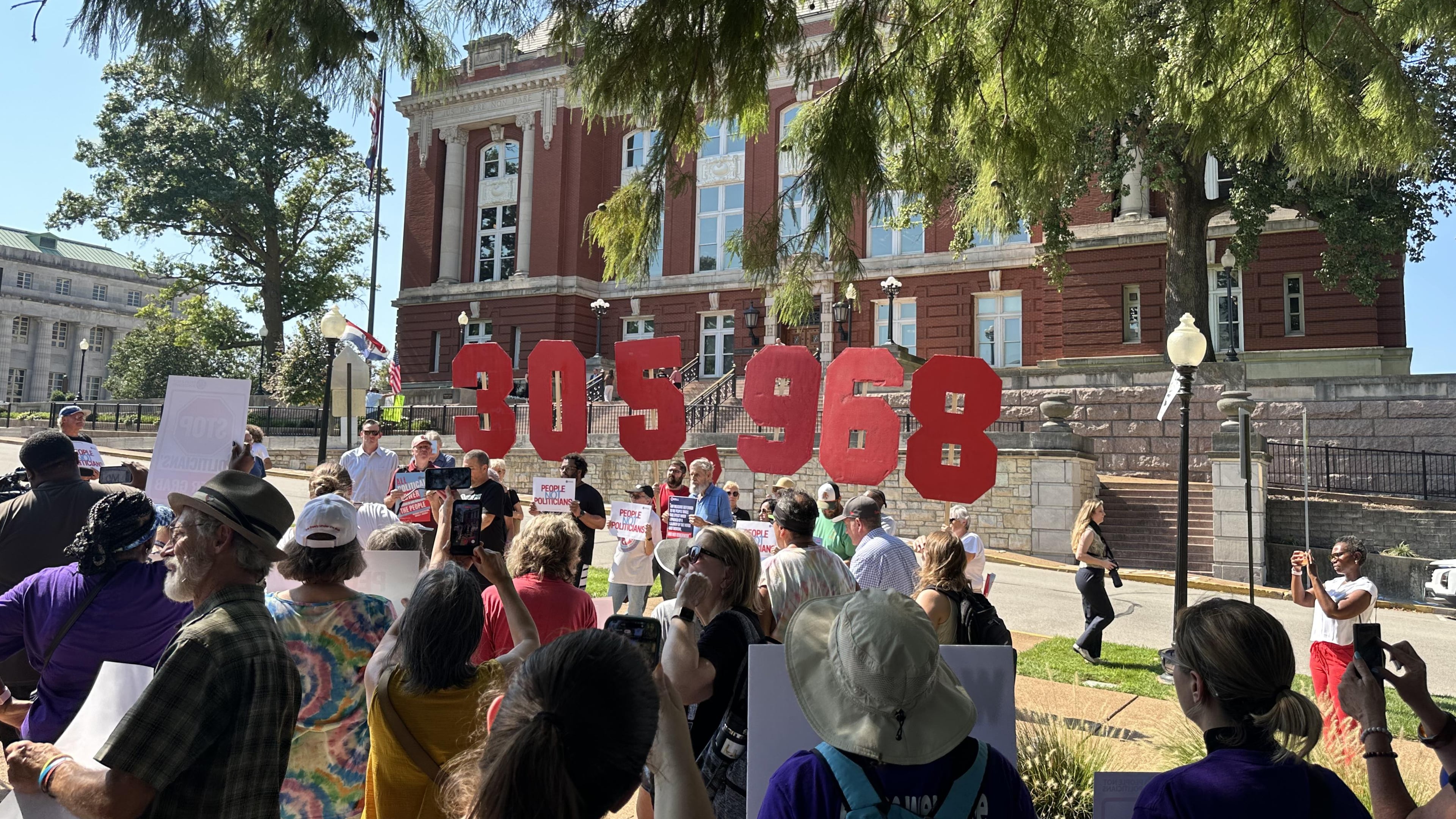 In this photo taken with a smartphone, people opposing a congressional redistricting plan rally outside the Missouri Supreme Court in Jefferson City, Mo., Wednesday, Sept. 2, 2026, while displaying the number of petition signatures submitted seeking a statewide vote on the measure. (AP Photo/David A. Lieb)