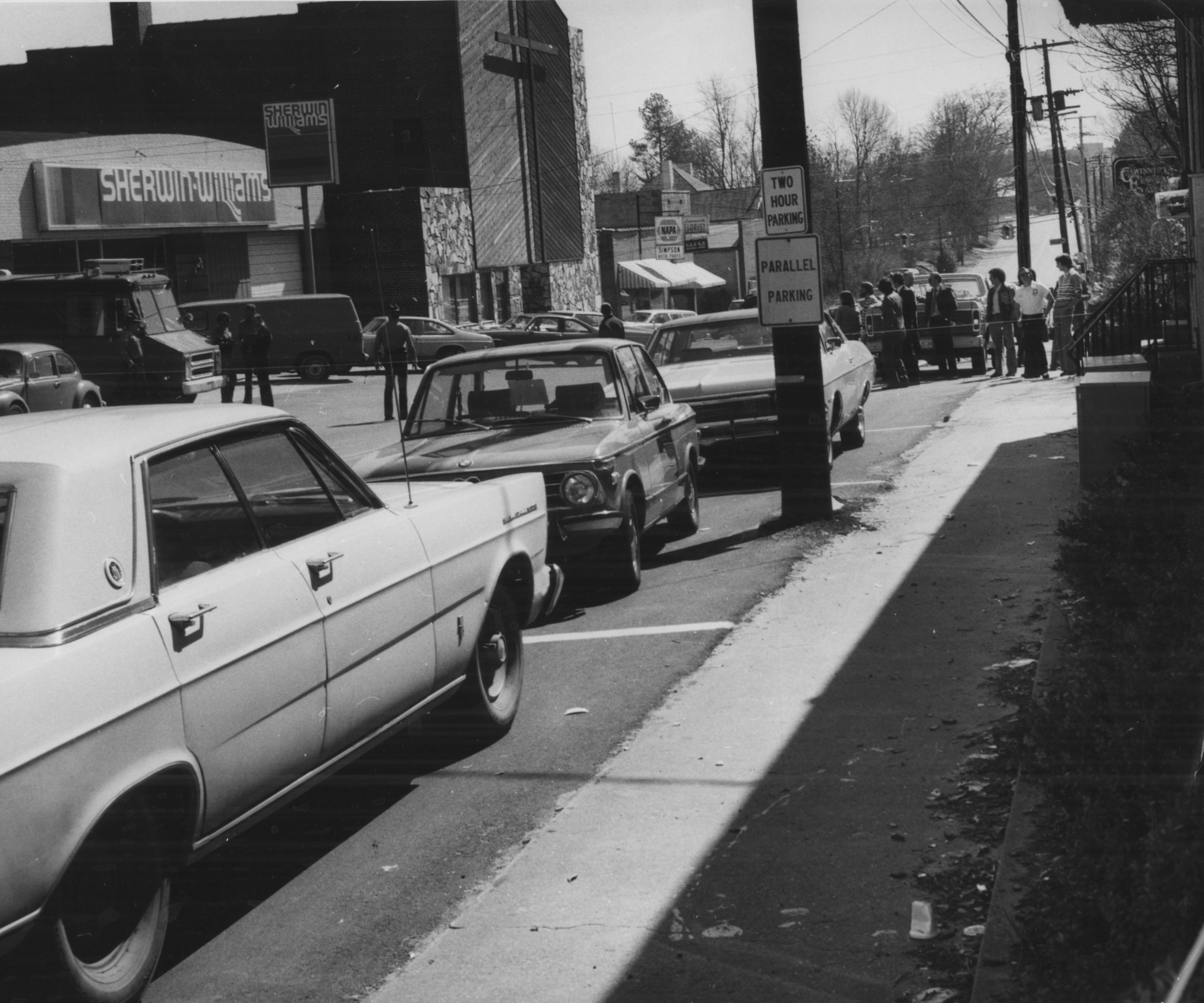Blood is visible on the sidewalk near the area where Flynt and Reeves fell after being hit. According to the photographer's caption, the men fell in the "area approximately where the third car is parked behind the parking signs." The Constitution staff report said that "in the confusion of the first few minutes, Gwinnett police issued a bulletin for a black and silver car, but then later said that car was not involved in the shooting."