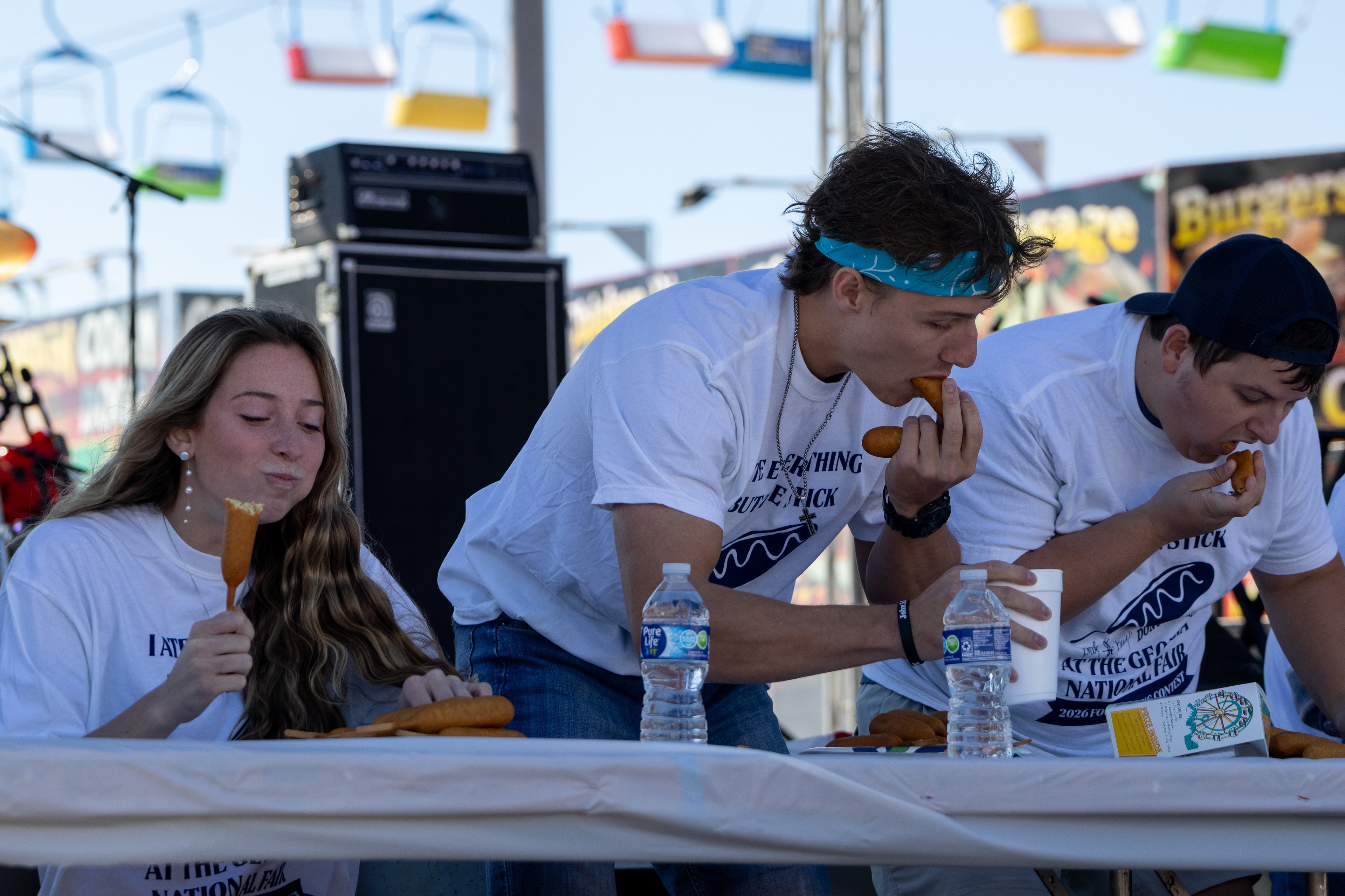 Alivia Blaton (left) chews on her corn dog next to her boyfriend Jesse Dean (middle) and competitor Cameron Perkins (right) at the corn dog eating competition at the 37th annual Georgia National Fair on Saturday, October 17, 2026. Dean won last year's hot dog eating competition after Blanton signed him up unknowingly, so she decided to participate this year while he defended his crown. Dean won first place back-to-back years, and Perkins won second. (Wesley Webb/UGA Photojournalism Workshop)