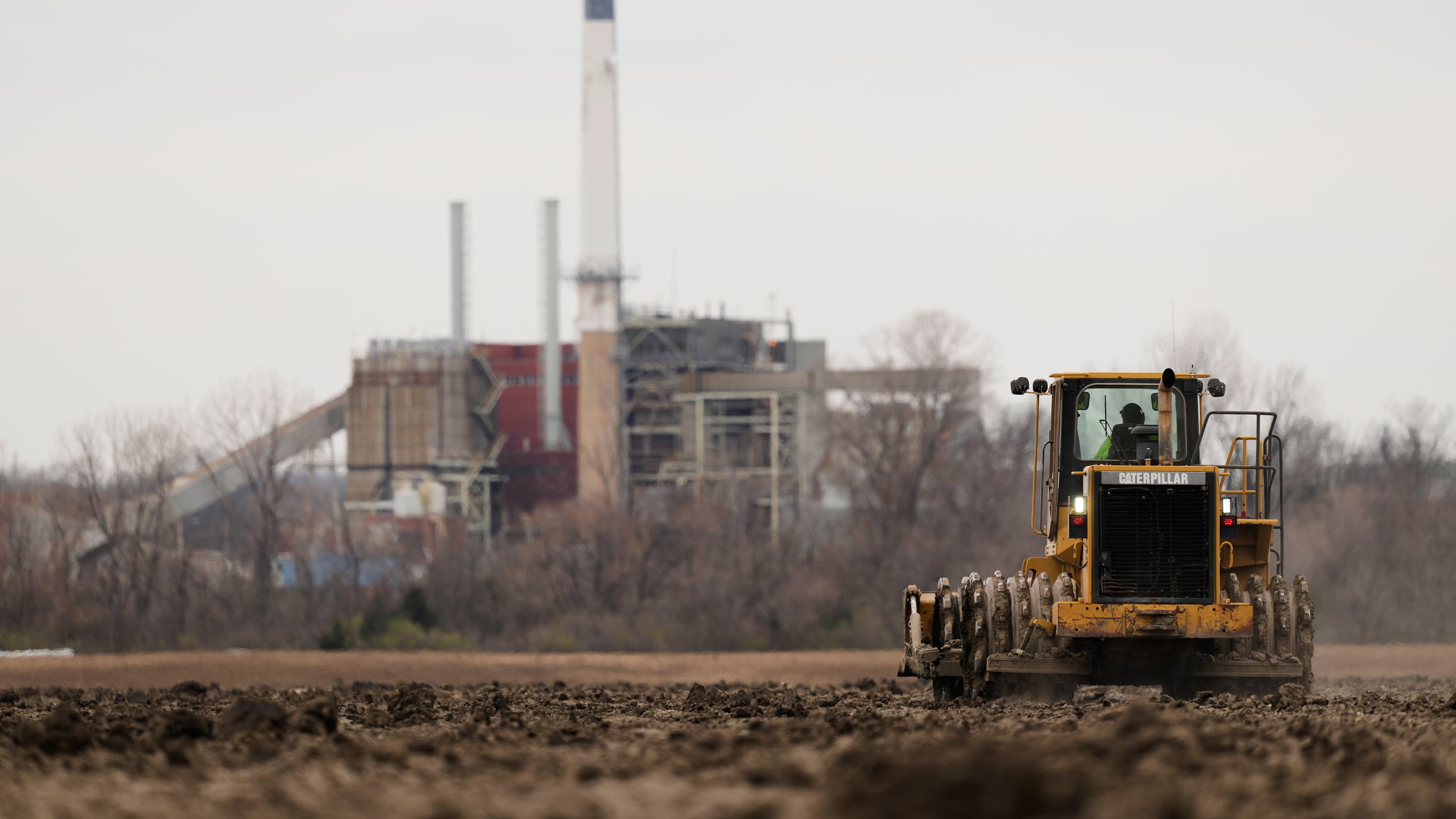 A worker prepares a plot of land for an AI data center, with a retired power plant being refurbished to provide electricity for the facility in the distance, March 24, 2026, in Independence, Mo. (Charlie Riedel/AP)
