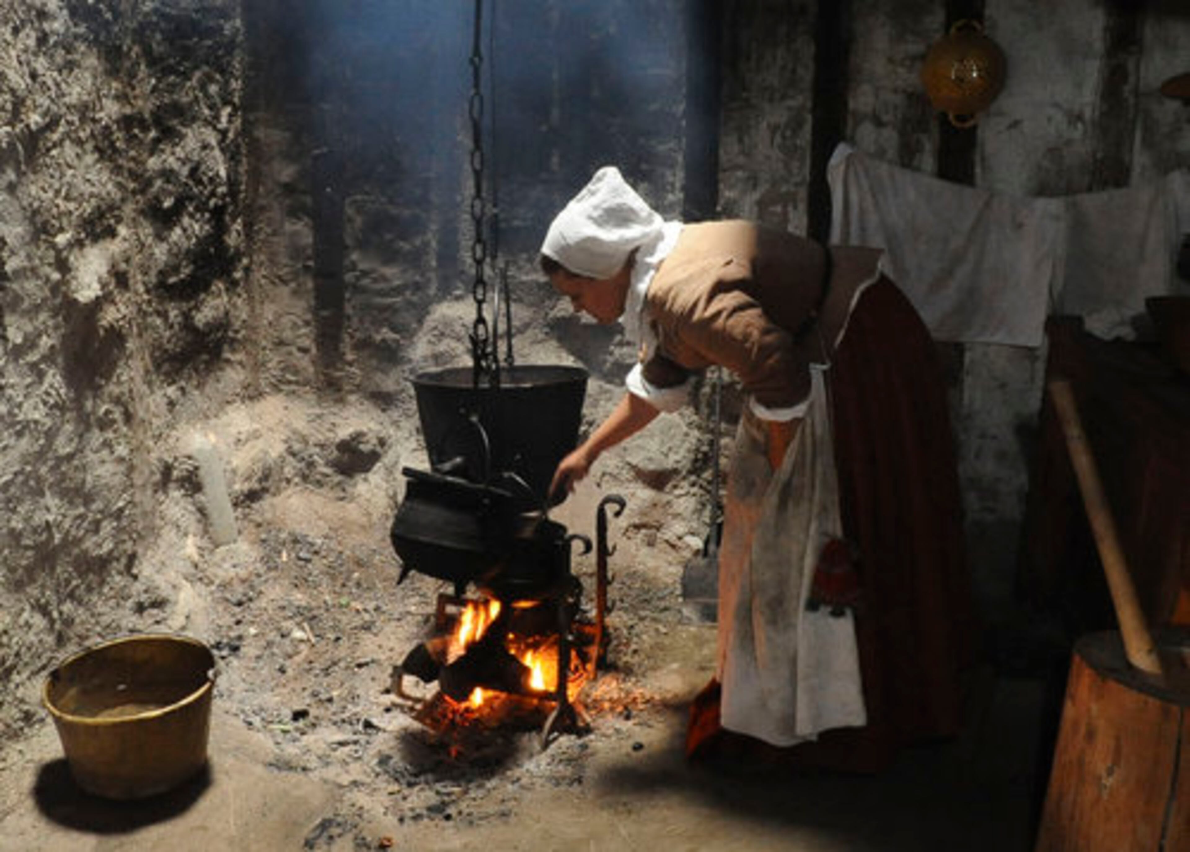 An employee of Plimoth Plantation portraying Priscilla Alden cooks hasty pudding over a fire in a timber-frame house.
