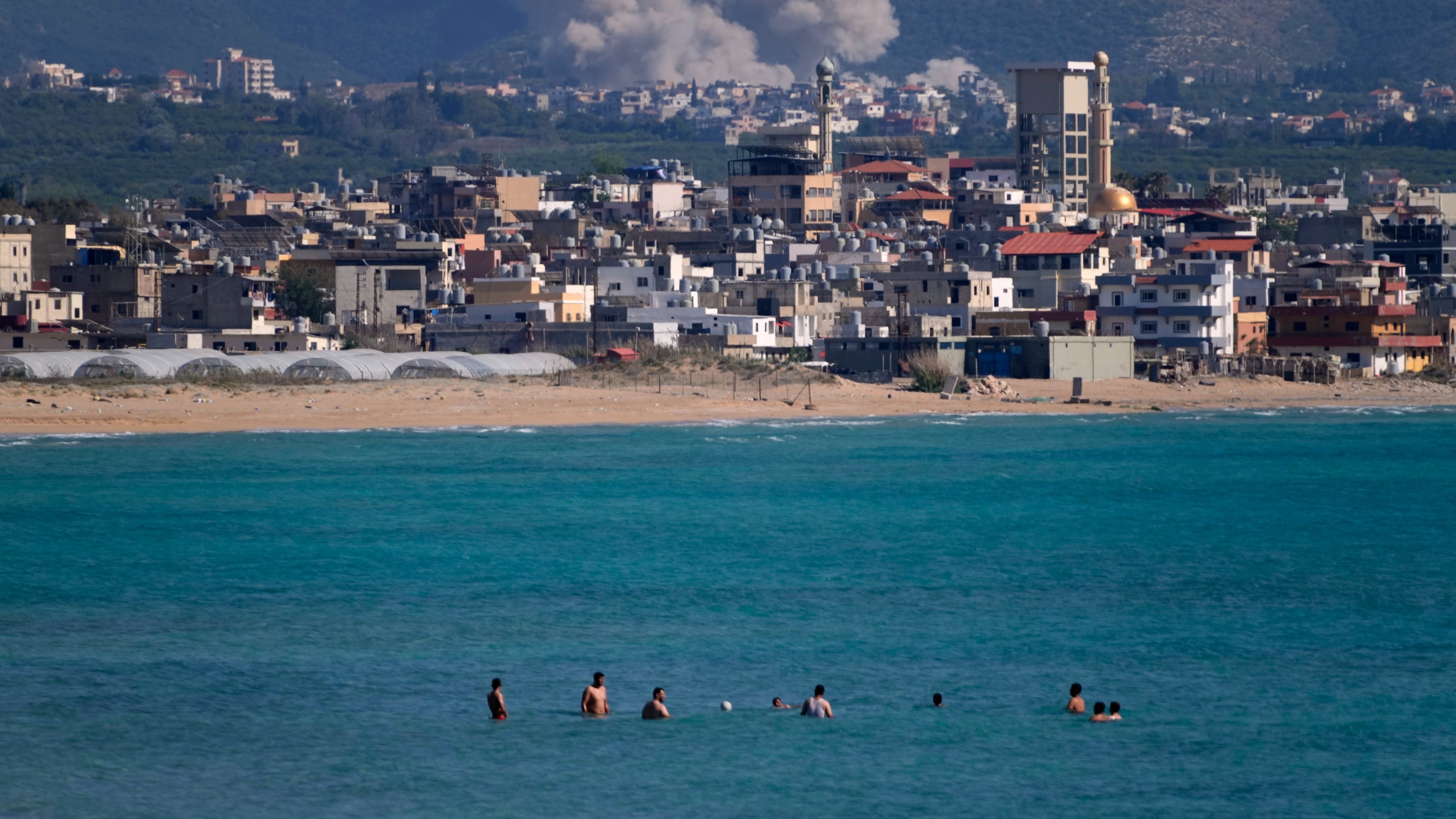 People swim on a public beach as smoke, background, rises from an Israeli airstrike that hit the Qlaileh village, seen from the southern port city of Tyre, Lebanon, Tuesday, June 2, 2026. (AP Photo/Mohammed Zaatari)