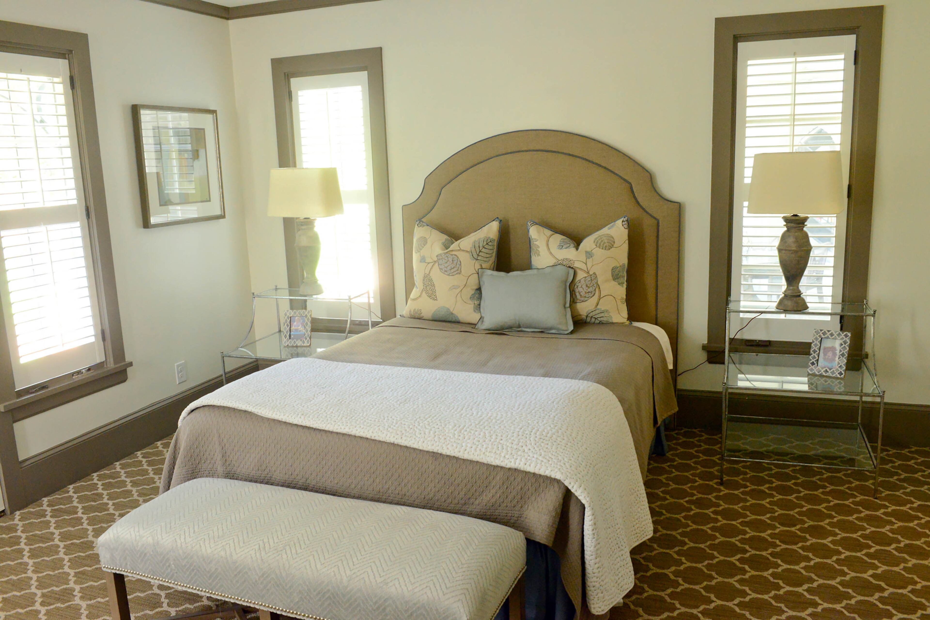 The guest bedroom in the basement benefits from the lot, with light coming through multiple windows. The upholstered headboard is from Peridot. The glass and metal side tables from Ballard Designs have elegant curved arms. The walls are painted Benjamin Moore's White Dove.