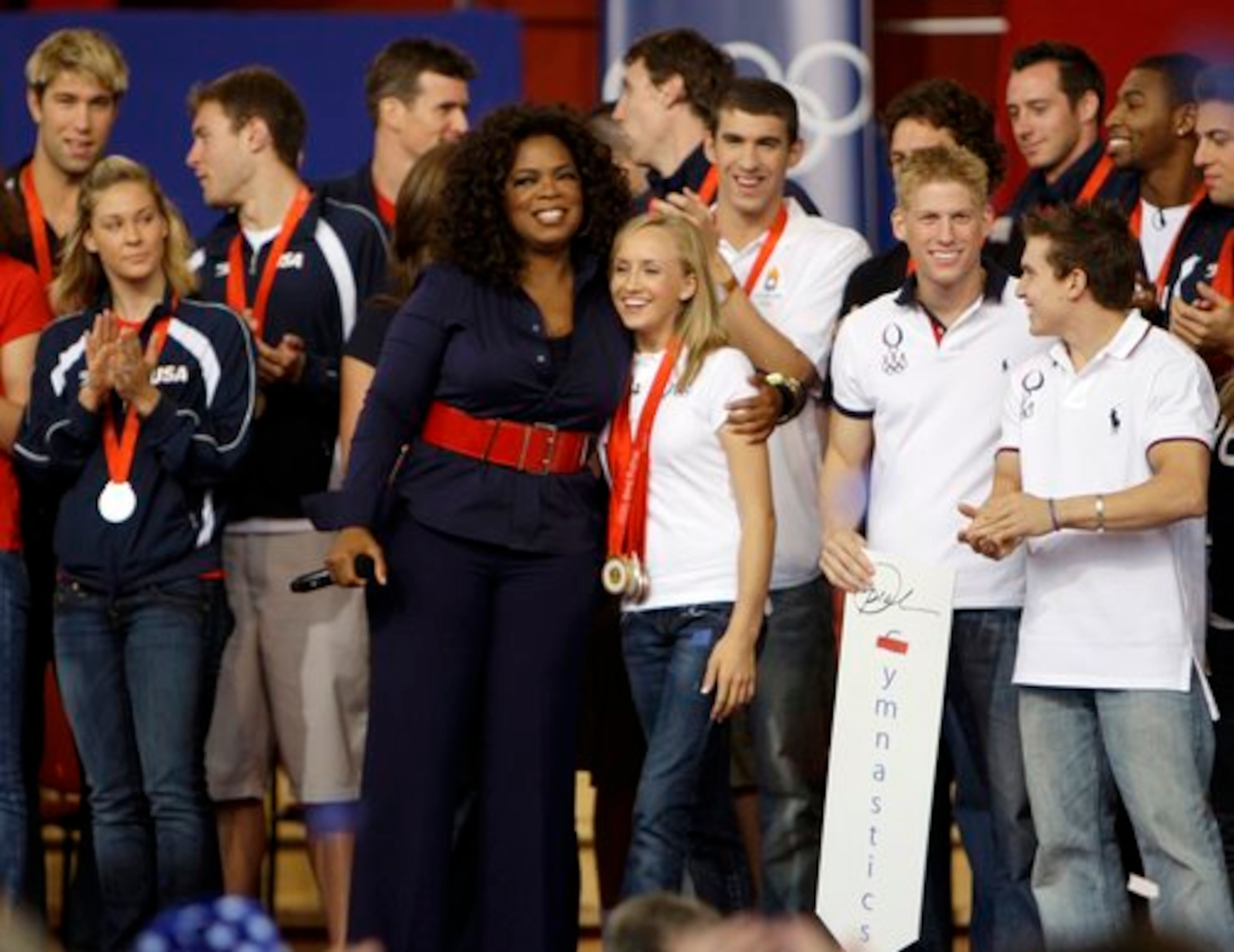 Oprah Winfrey hugs Olympic gold medalist Nastia Liukin as gold medalist Michael Phelps, behind Liukin, applauds.
