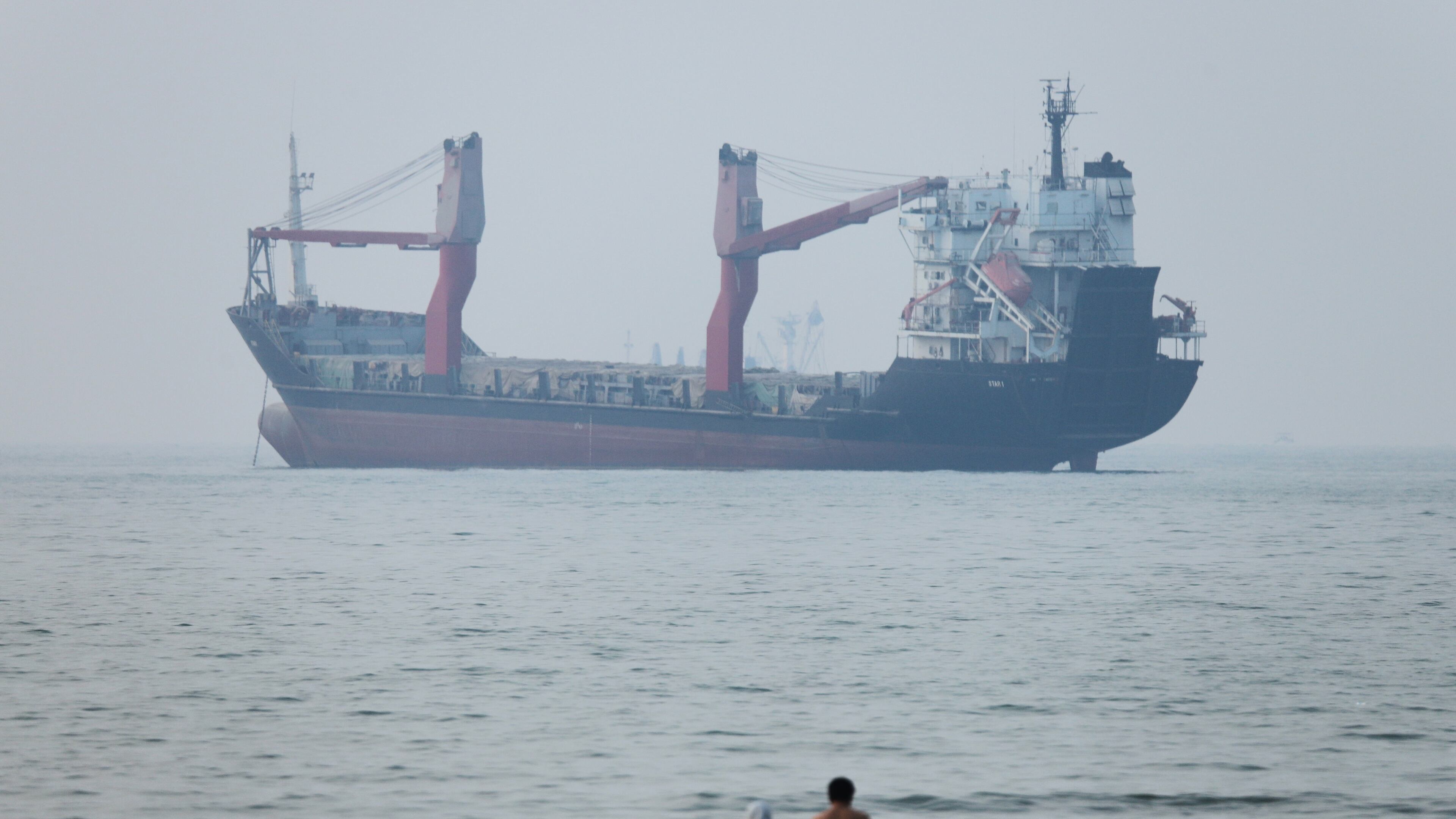 A group of people stands in shallow water as a cargo ship appears anchored in the Strait of Hormuz off Bandar Abbas, Iran, Tuesday, June 30, 2026. (Amirhosein Khorgooi/ISNA via AP)