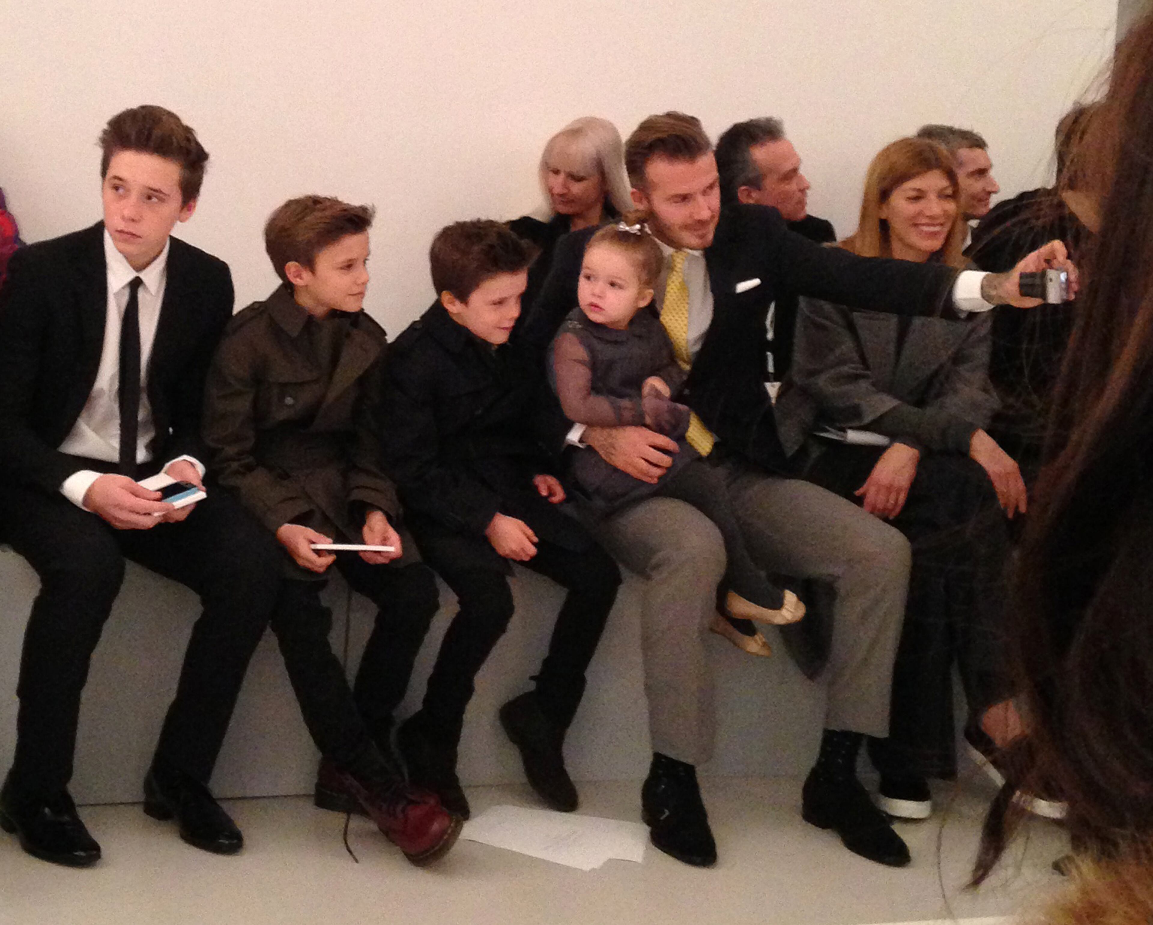 David Beckham takes a photo of his four children in the front row before the Victoria Beckham show, as daughter Harper sits in Beckham's lap Sunday Feb. 9, 2014, during Fashion Week in New York. (AP Photo/Jocelyn Noveck)