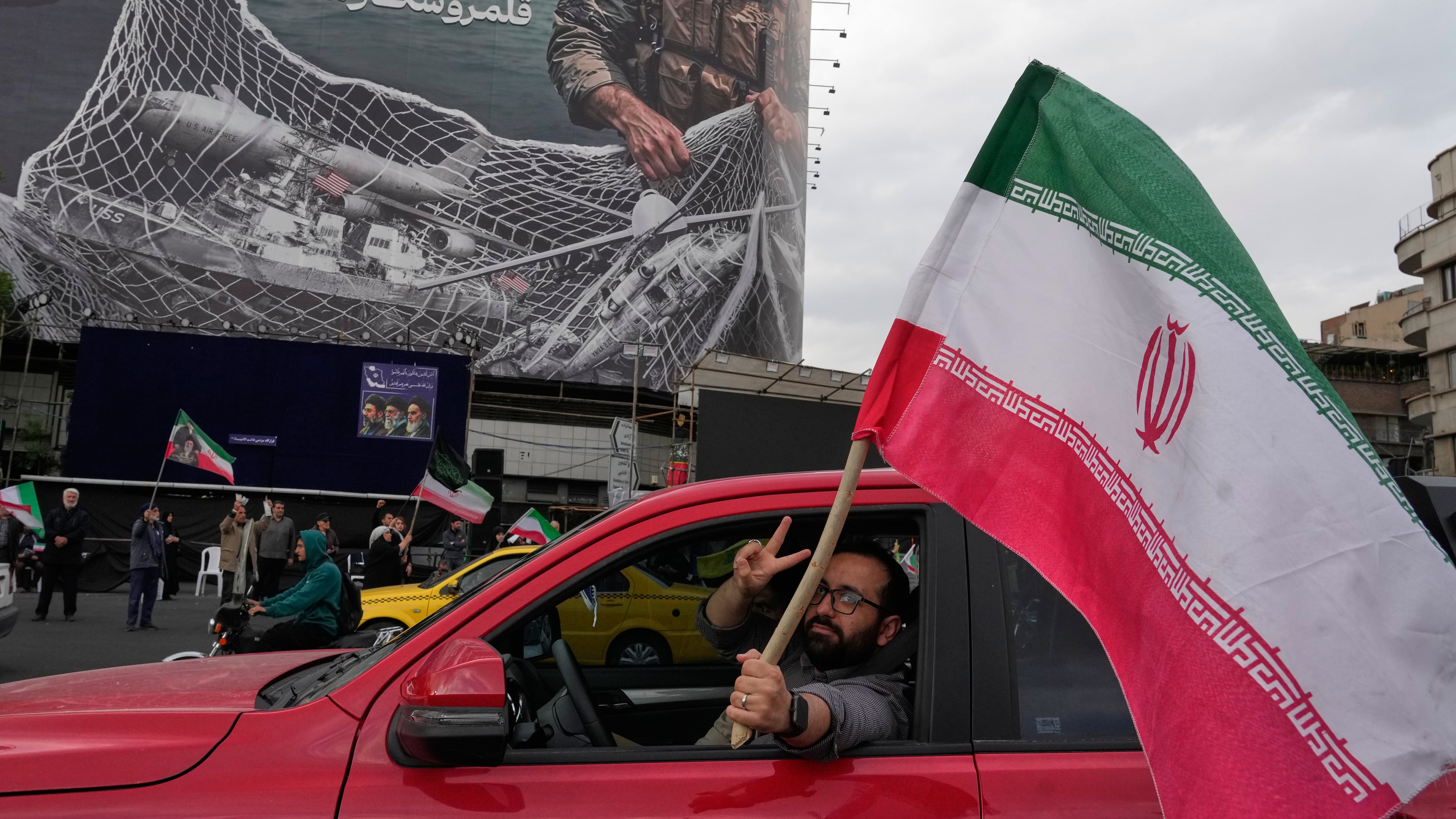 A man flashes a victory sign as he carries an Iranian flag in front of an anti-U.S. billboard depicting the American aircrafts into the Iranian armed forces fishing net with signs that read in Farsi: "The Strait of Hormuz will remain closed, The entire Persian Gulf is our hunting ground," at the Eqelab-e-Eslami, or Islamic Revolution Square in downtown Tehran, Iran, Monday, April 13, 2026. (AP Photo/Vahid Salemi)