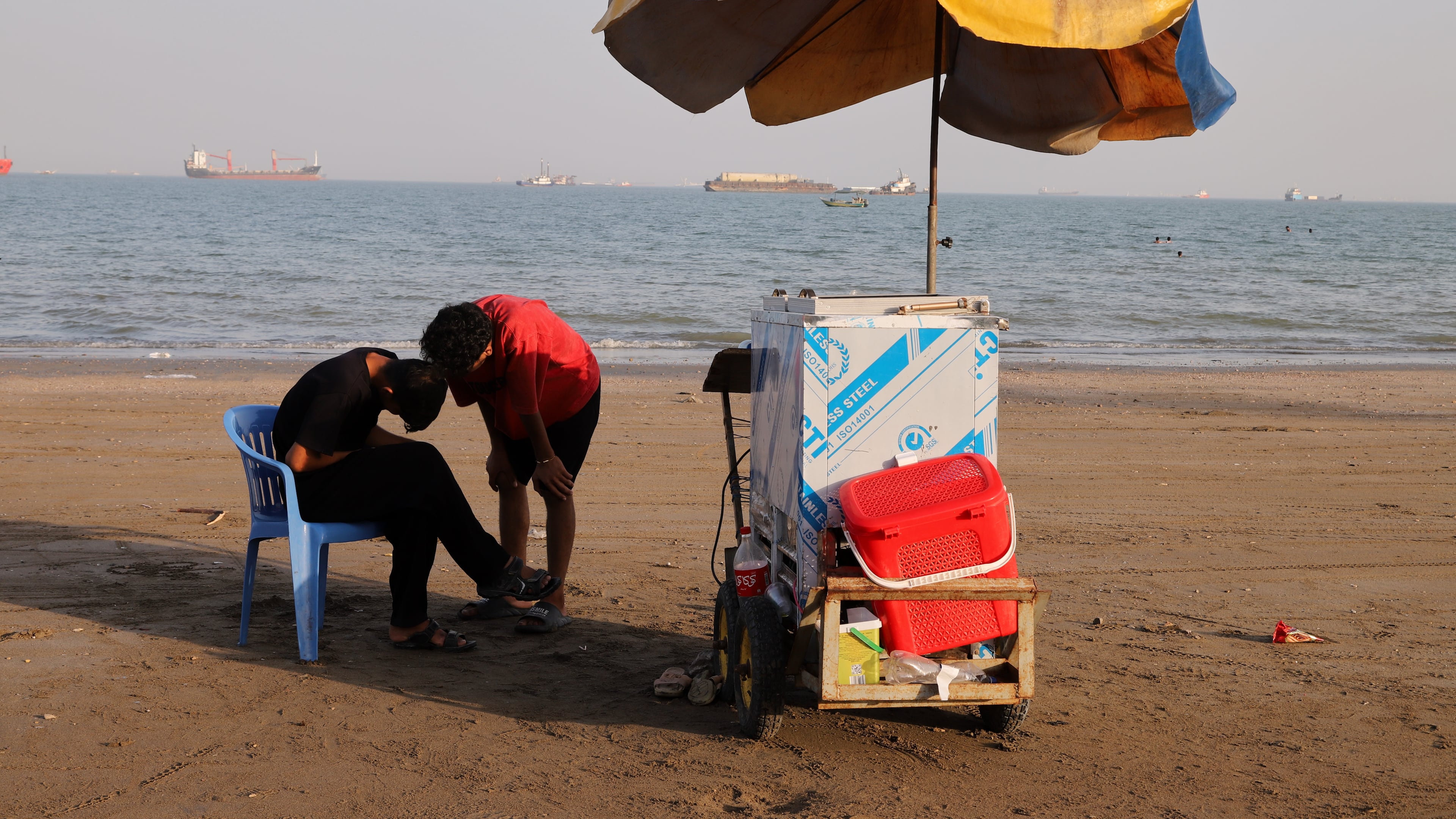 People stand next to a vendor's cart on the beach as cargo ships and offshore service vessels are anchored in the Strait of Hormuz, off Bandar Abbas, Iran, Sunday, May 31, 2026. (Amirhosein Khorgooi/ISNA via AP)