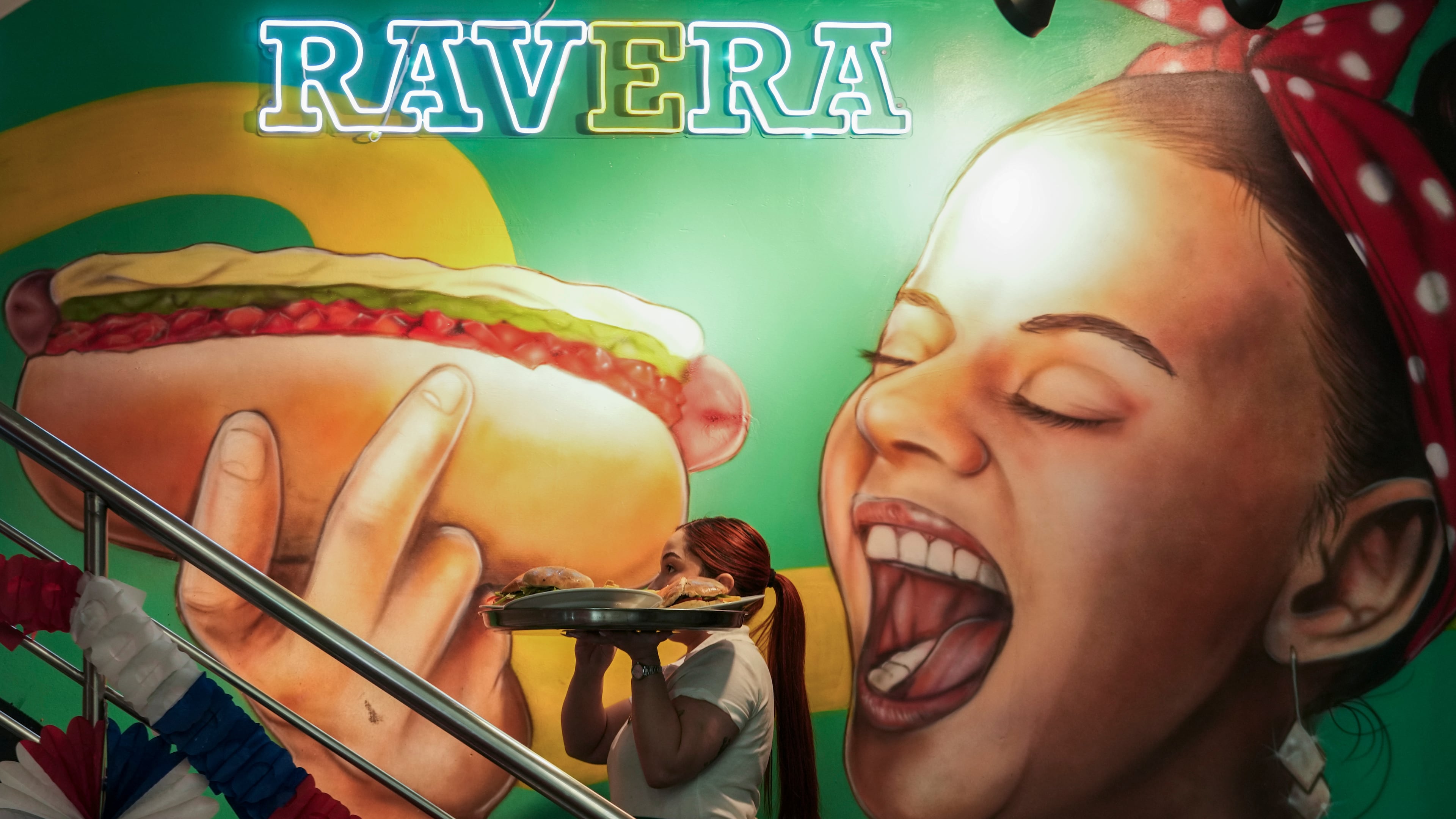 A waitress carries a tray with "completos", traditional Chilean hot dogs topped with mashed avocado, diced tomatoes and mayonnaise, past a mural dedicated to the dish at the Ravera restaurant in downtown Santiago, Chile, Thursday, Sept. 10, 2026. (AP Photo/Esteban Felix)