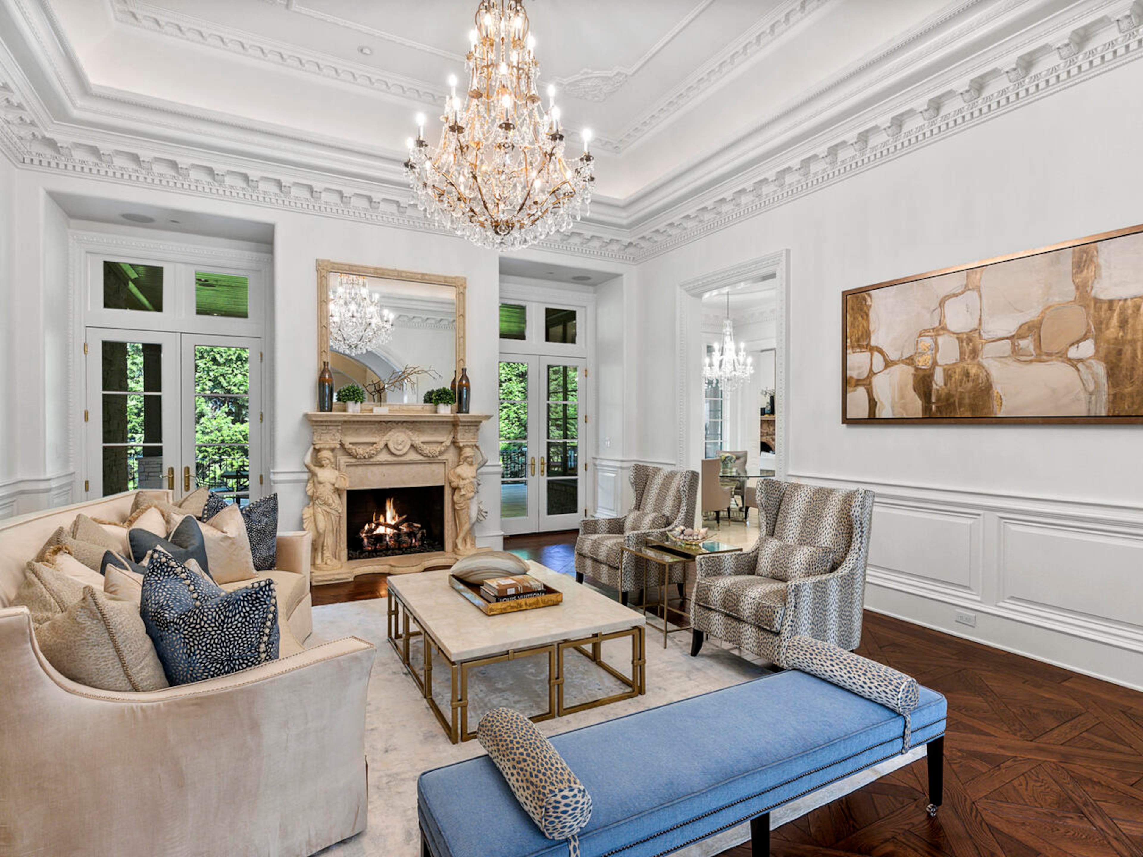 Intricate details adorn the formal living room – from the wainscotting, to the incredibly detailed molding and tray ceiling with the centered Waterford chandelier hanging in the center of the room, to the marble fireplace surrounds flanked by Greek statues. French doors flank the fireplace leading to the outdoor loggia. Text by Elizabeth Manderson and Julie Pedersen/Photo by Ilya Zobanov, Gold Lens Media.