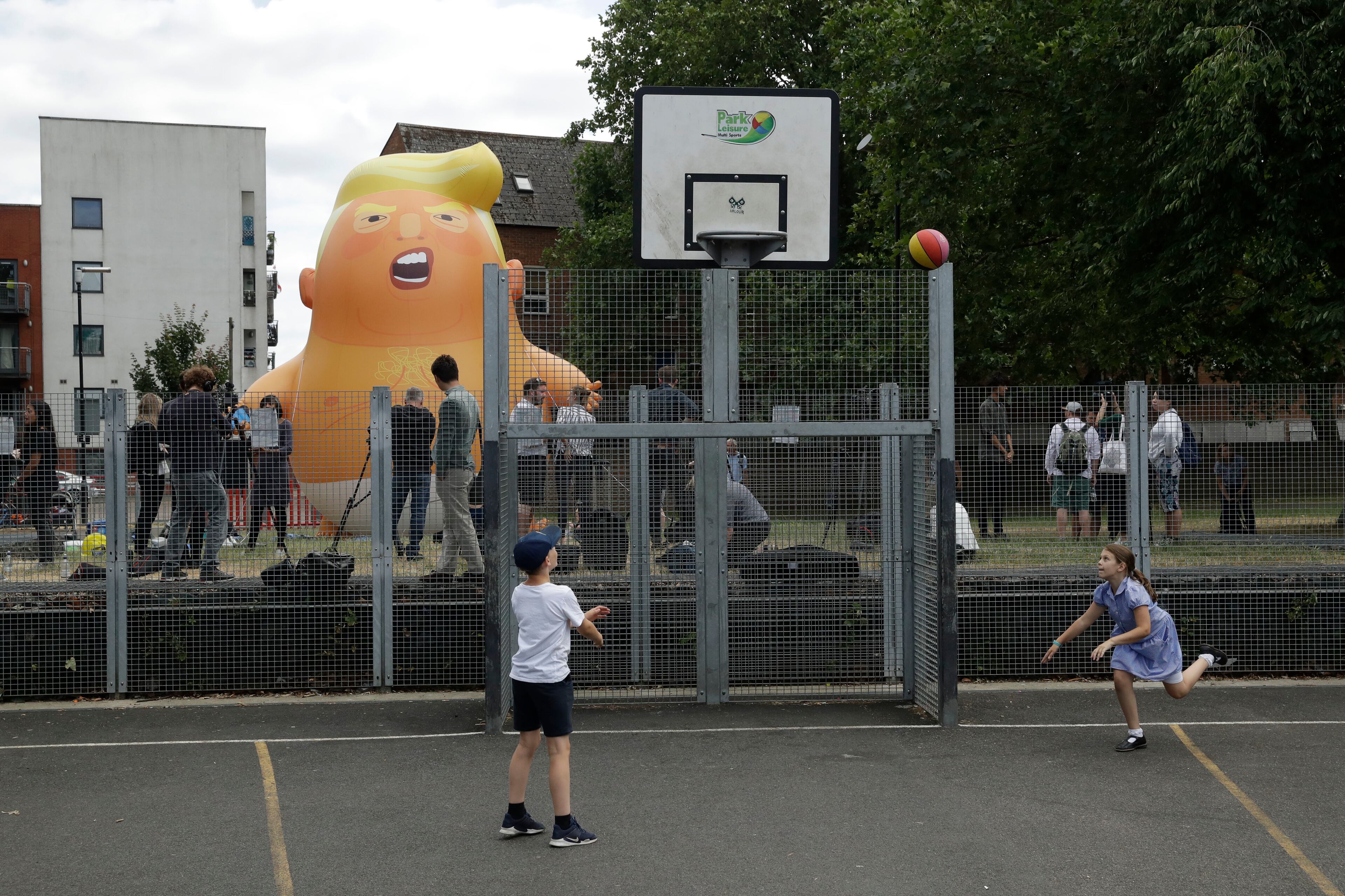 In this photo taken on Tuesday, July 10, 2018, children play basketball in the foreground as a six-meter high cartoon baby blimp of U.S. President Donald Trump stands inflated during a practice session in Bingfield Park, north London. Trump will get the red carpet treatment on his brief visit to England that begins Thursday: Military bands at a gala dinner, lunch with the prime minister at her country place, then tea with the queen at Windsor Castle before flying off to one of his golf clubs in Scotland. But trip planners may go out of their way to shield Trump from viewing another aspect of the greeting: an oversize balloon depicting the president as an angry baby in a diaper that will be flown from Parliament Square during what are expected to be massive gatherings of protesters opposed to Trumpâs presence. (AP Photo/Matt Dunham)