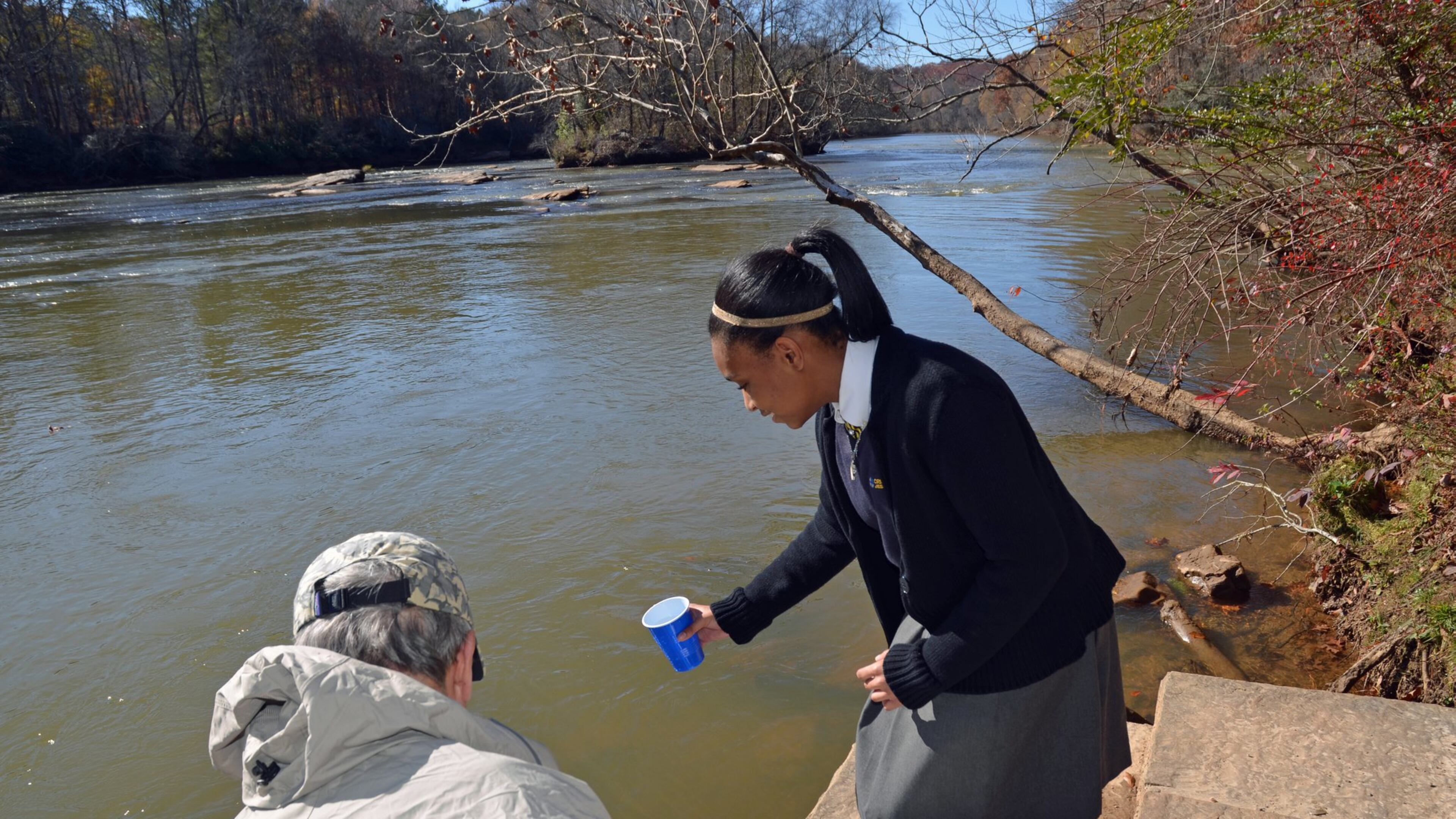 Cristo Rey science student Jasmine Mitchell assists Jim Byrd of the Upper Chattahoochee chapter of Trout Unlimited release trout into the river near Peachtree Corners. Students at the Midtown school raised about 250 trout to help the organization’s conservation efforts. Photo credit: Sylvia Small.