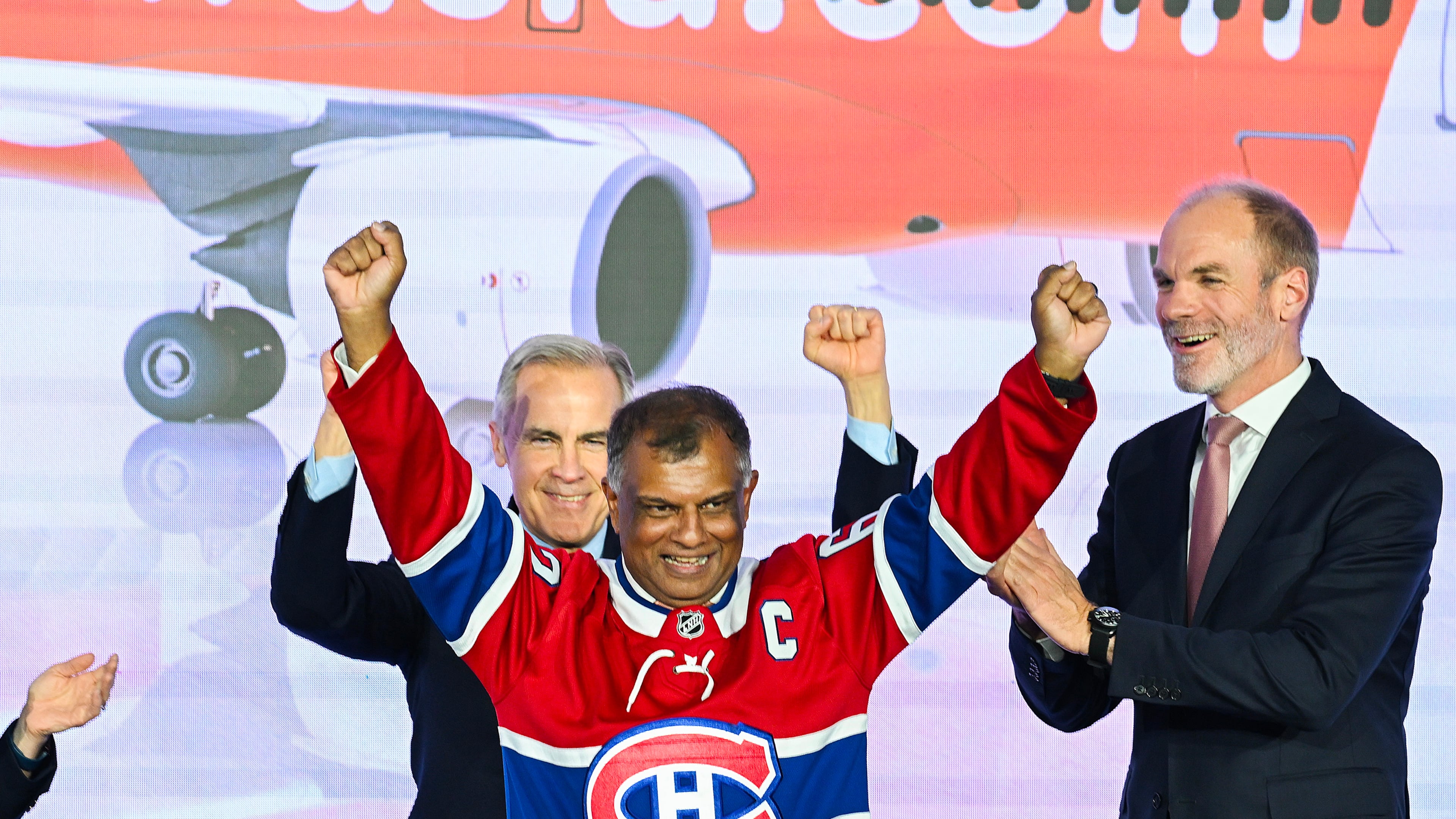Canada's Prime Minister Mark Carney, left, and Lars Wagner, CEO, Airbus Commercial Aircraft, right, cheer on AirAsia CEO Tony Fernandes, centre, during an event at the Airbus A220 Delivery Center in Mirabel, Que., Wednesday, May 6, 2026, where AirAsia placed a landmark order for 150 Airbus A220 aircraft. (Graham Hughes/The Canadian Press via AP)