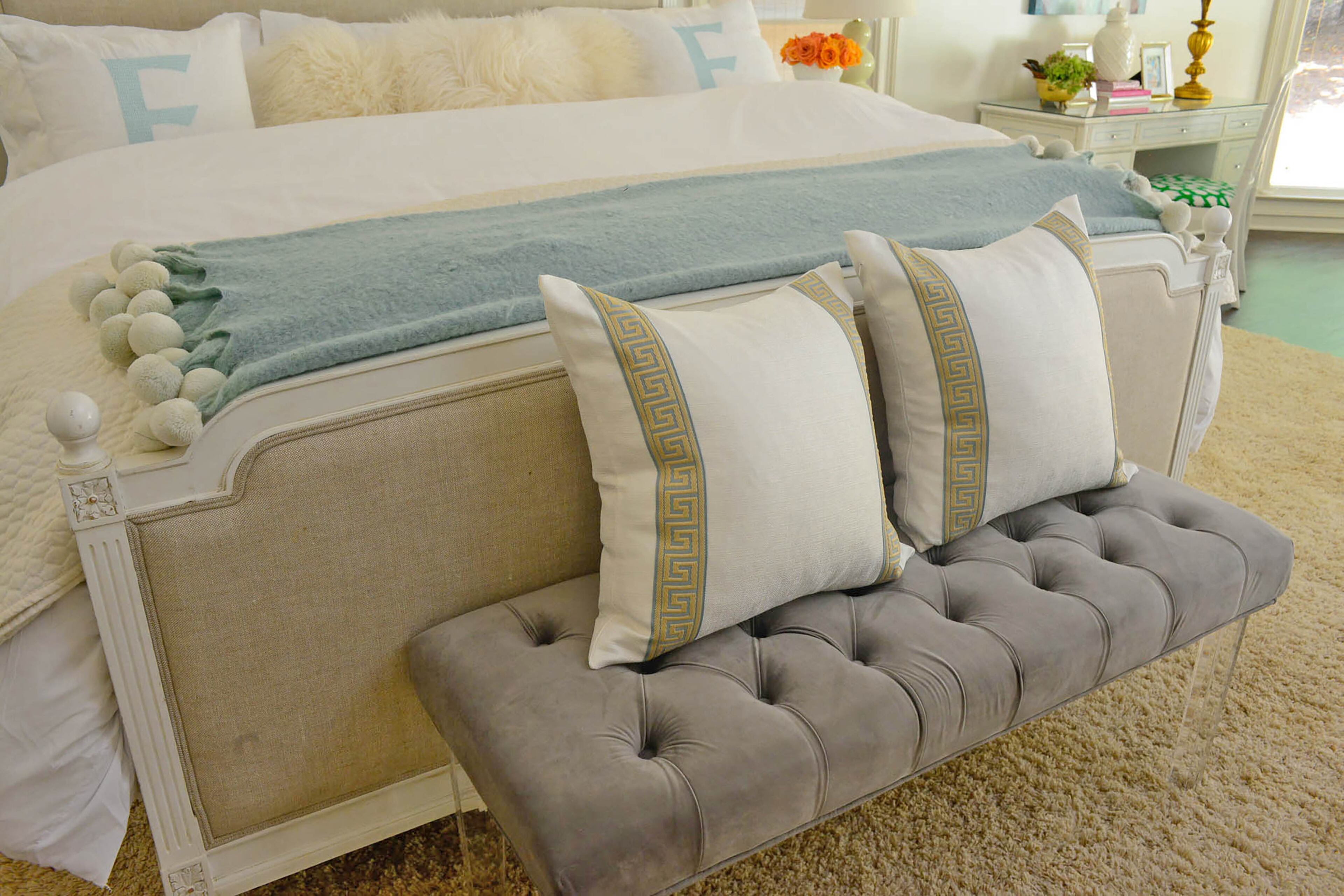 Calm neutrals fill homeowner the master bedroom. Monogrammed pillows and a throw blanket add subtle hues of blue to the upholstered bed from RH, which is flanked by vintage mirrored nightstands. A tufted bench from Wayfair with clear acrylic legs plays into the timeless style of the space.