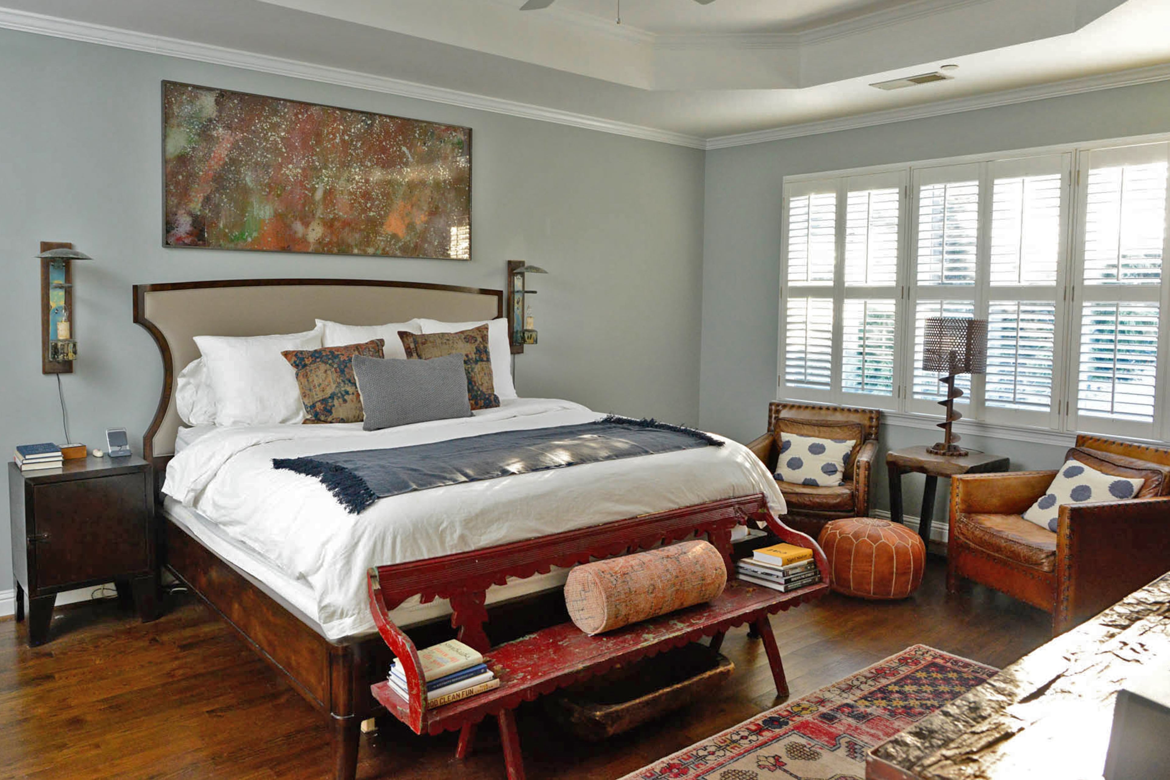 A wood upholstered bed, with its unusual headboard shape, is from Scott Antique Markets. A pair of nightstands by Industrial Chic Loft flank it with lights from Kudzu Antiques. The art piece above the bed is a painted mirror by Timothy Poe. The pair of leather chairs and auger lamp are from Scott Antique Markets.