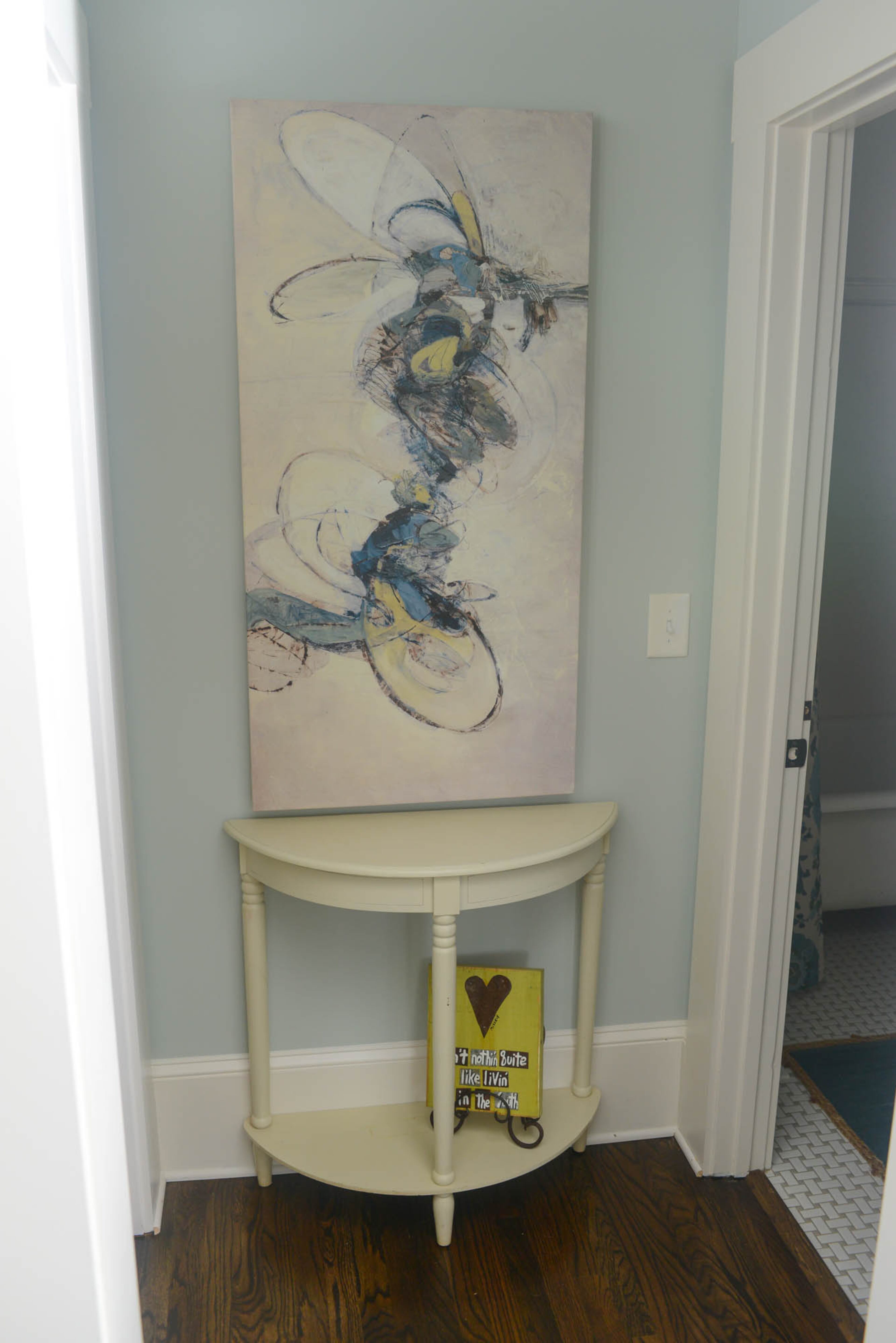 Dark-stained hardwood flooring contrasts with the pale blue-green walls in the hallway and a matte gray dot porcelain basketweave tile floor in the adjacent guest bath. A simple cream-colored demilune accent table provides balance for the artwork with a strong sense of movement hanging above.