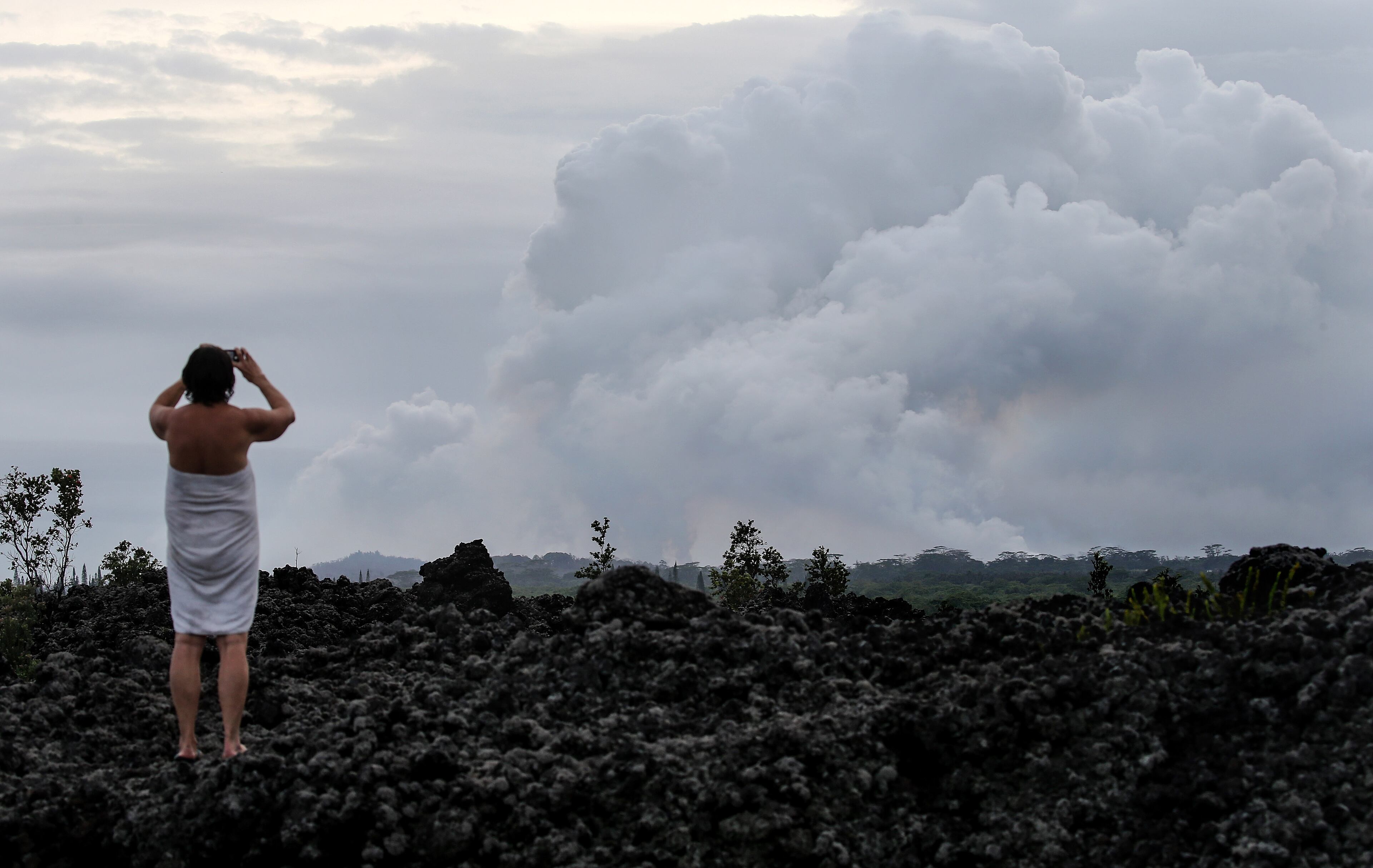 PAHOA, HI - MAY 10: A local resident stands on an old lava flow while taking pictures of a plume of volcanic gas mixed with smoke from fires caused by lava rising from the vicinity of the Leilani Estates neighborhood, in the aftermath of eruptions and lava flows from the Kilauea volcano on Hawaii's Big Island, on May 10, 2018 in Pahoa, Hawaii. The U.S. Geological Survey said a recent lowering of the lava lake at the volcano's Halemaumau crater has raised the potential for explosive eruptions at the volcano. Vog, a haze or smog containing gases, smoke and dust from volcanic eruptions, may eventually spread from the eruptions to other islands in Hawaii. (Photo by Mario Tama/Getty Images)