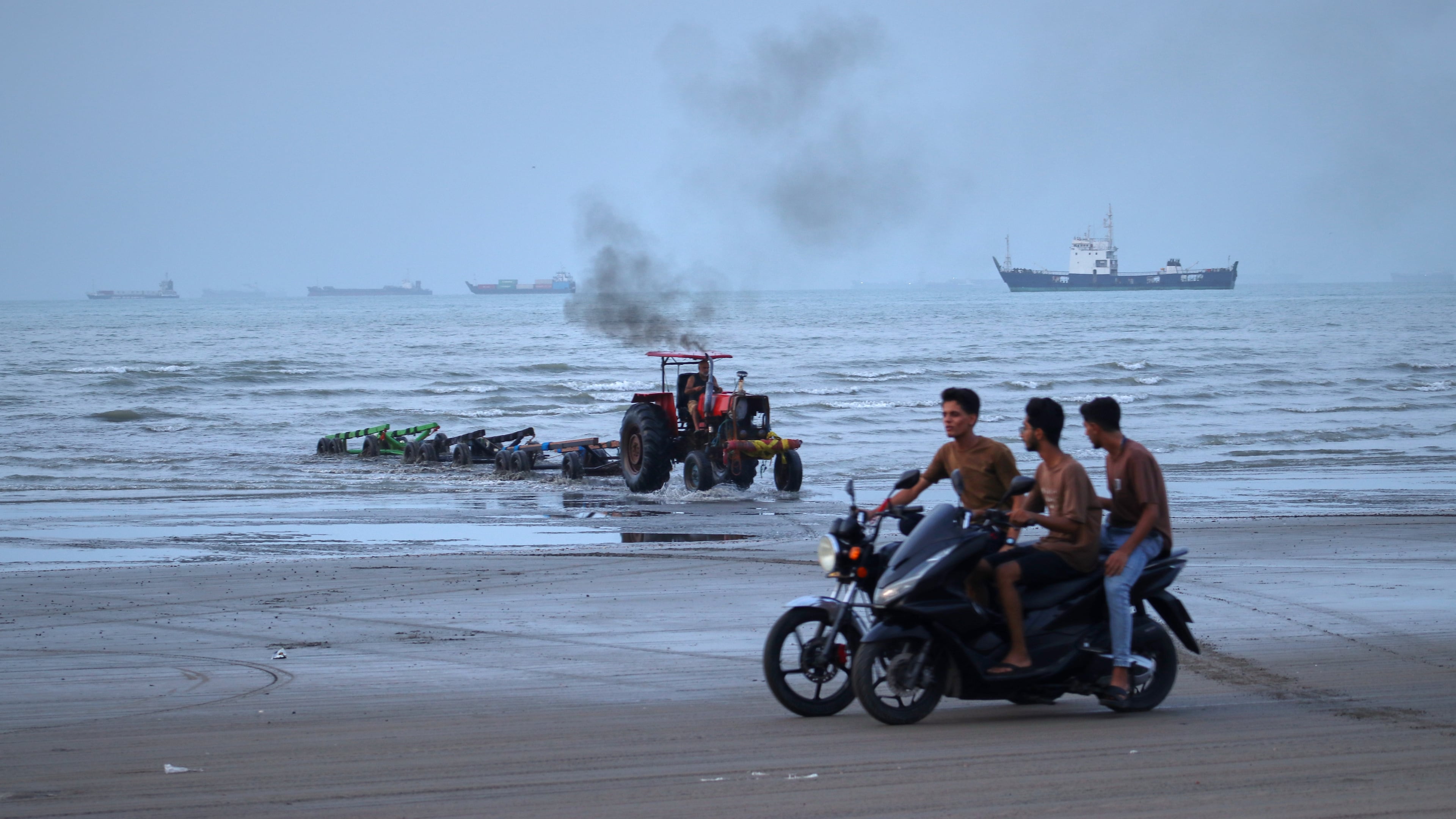 Young men ride motorcycles along the shore as a tractor pulls equipment from the water and commercial vessels appear anchored in the Strait of Hormuz off Bandar Abbas, Iran, Monday, July 27, 2026. (Razieh Poudat/ISNA via AP)