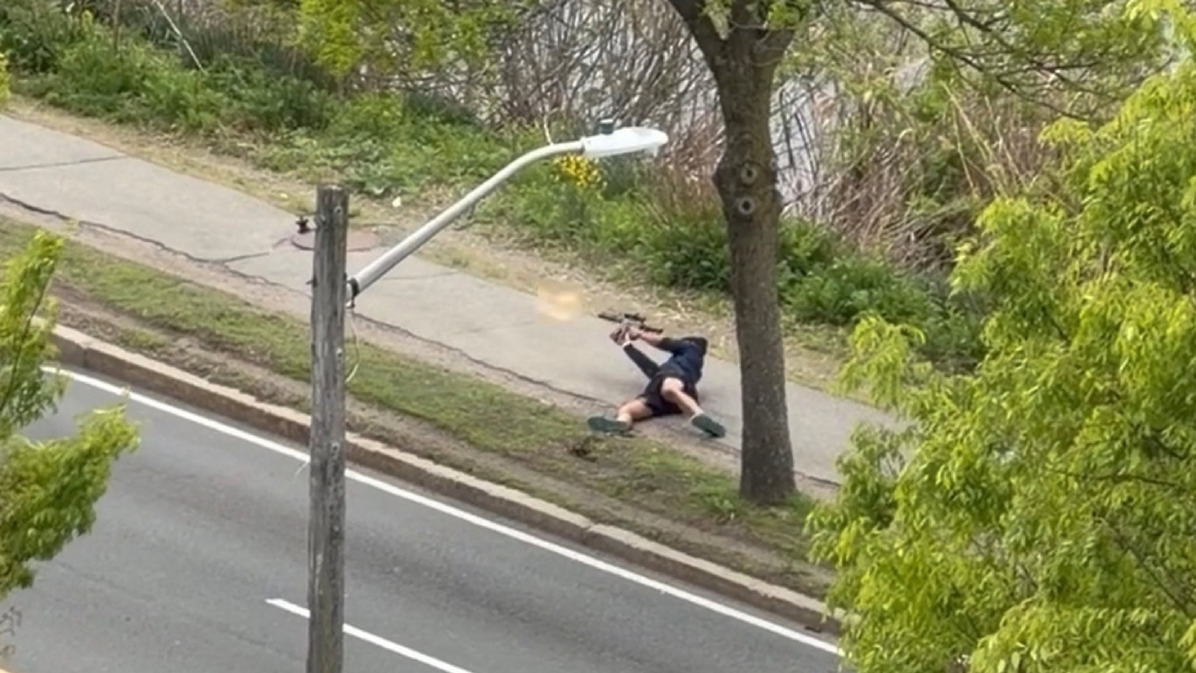 This image taken from video provided by Youssef Adel, shows a man with an assault-style rifle firing his weapon at a busy road outside at in Cambridge, Mass. on Monday, May 11, 2026. (Youssef Adel via AP)