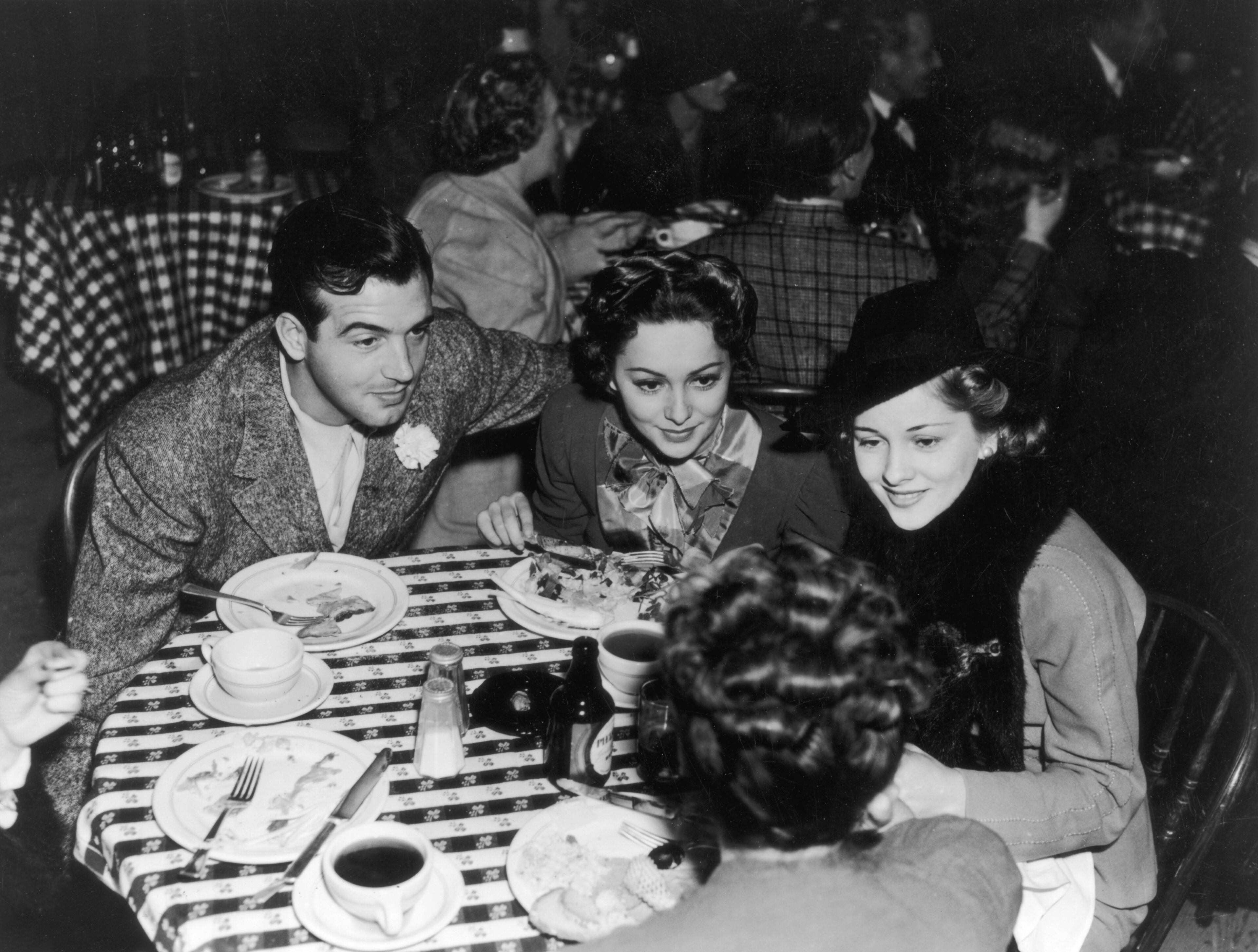 circa 1940: Olivia De Havilland leans in to hear a conversation while lunching with her sister Joan Fontaine and actor John Payne. (Photo by General Photographic Agency/Getty Images)