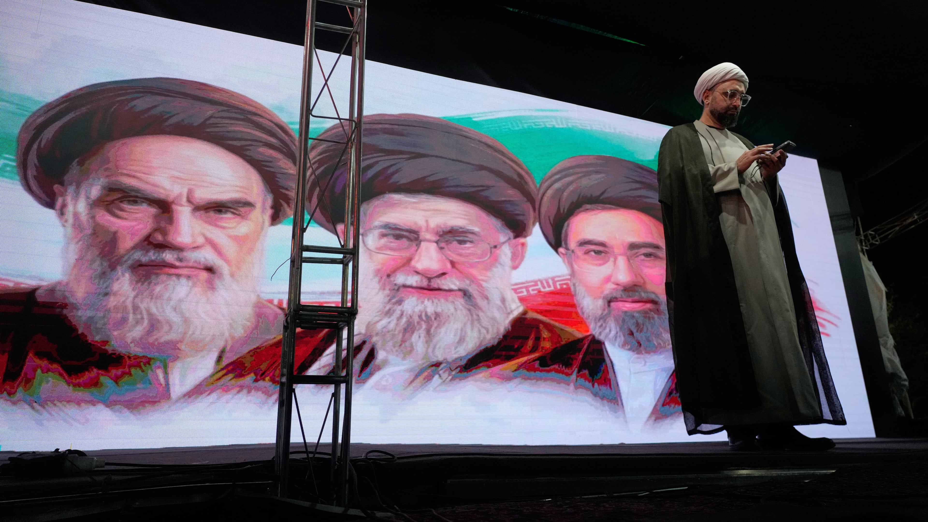 A cleric checks his cell phone on stage in front of a screen displaying portraits of the late Iranian revolutionary founder Ayatollah Khomeini, left, late Supreme Leader Ayatollah Ali Khamenei, center, and Supreme Leader Ayatollah Mojtaba Khamenei, during a pro-government gathering in Tehran, Iran, Sunday, June 7, 2026. (AP Photo/Vahid Salemi)
