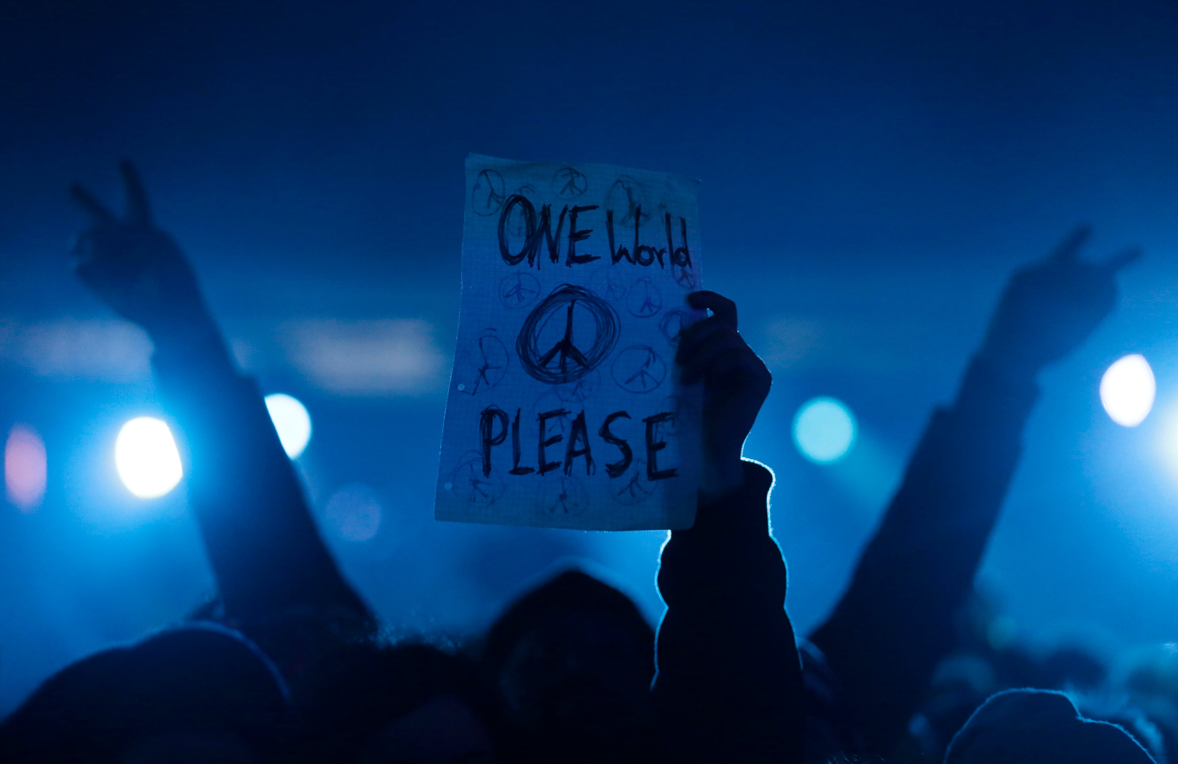 A person flashes the victory sign behind a man holding paper with a peace sign and the writing 'one world please' as they attend the central event to commemorate the Fall of the Wall at the Brandenburg Gate in Berlin, Germany, Sunday, Nov. 9, 2014. The 25th anniversary of the fall of the wall on Nov. 9, 1989 is marked with numerous events on the weekend. (AP Photo/Markus Schreiber)