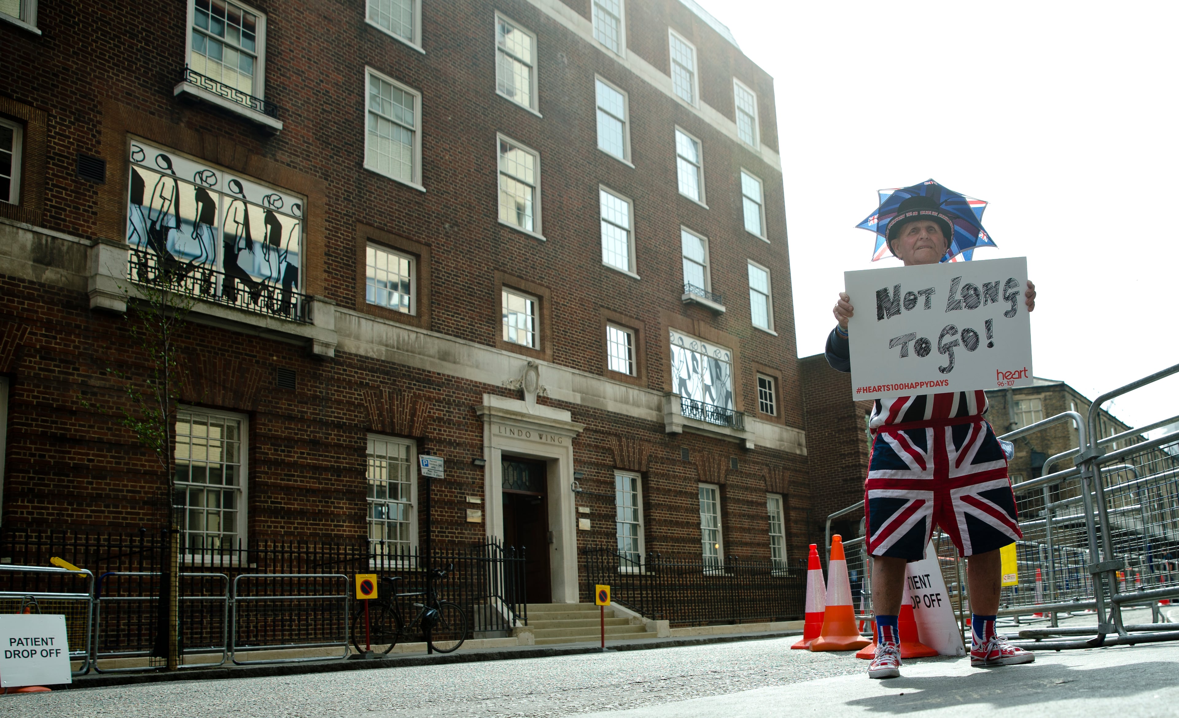 FILE - In this Friday, April 24, 2015 file photo, Terry Hutt poses for the media with a sign that reads 'Not Long to Go' as he waits with other royal fans, outside the Lindo wing at St Mary's Hospital in London. Kensington Palace says Prince William's wife, the Duchess of Cambridge has entered a London hospital to give birth to the couple's third child. The former Kate Middleton traveled by car on Monday, April 23, 2018 to the private Lindo Wing of St. Mary's Hospital in central London. The palace says she was in "the early stages of labor." (AP Photo/Alastair Grant, File)