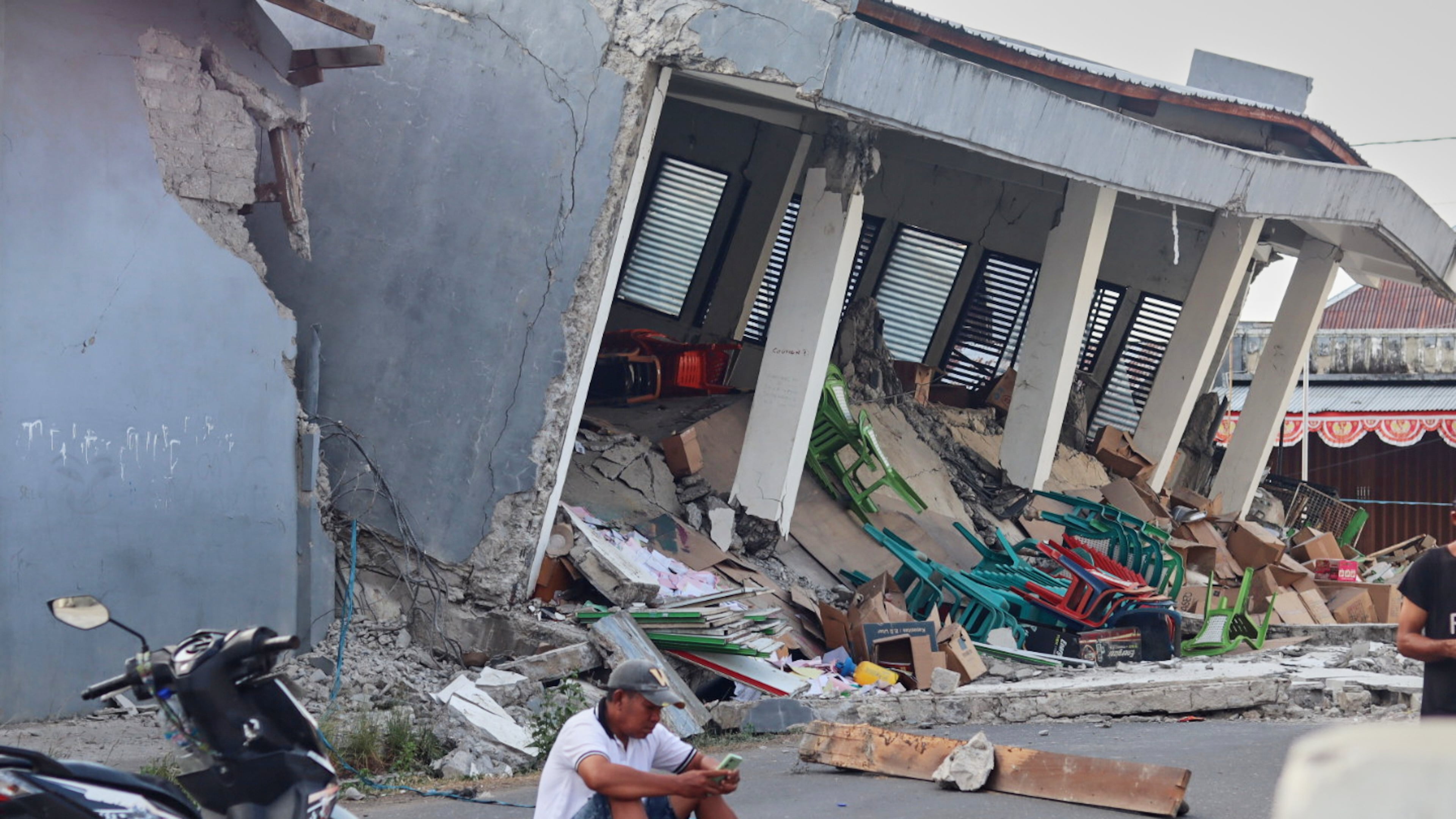 A man checks his phone near a building collapsed due to an earthquake, in Reok, East Nusa Tenggara province, Indonesia, Sunday, Aug. 16, 2026. (AP Photo/Aquila Handayani Abbas)