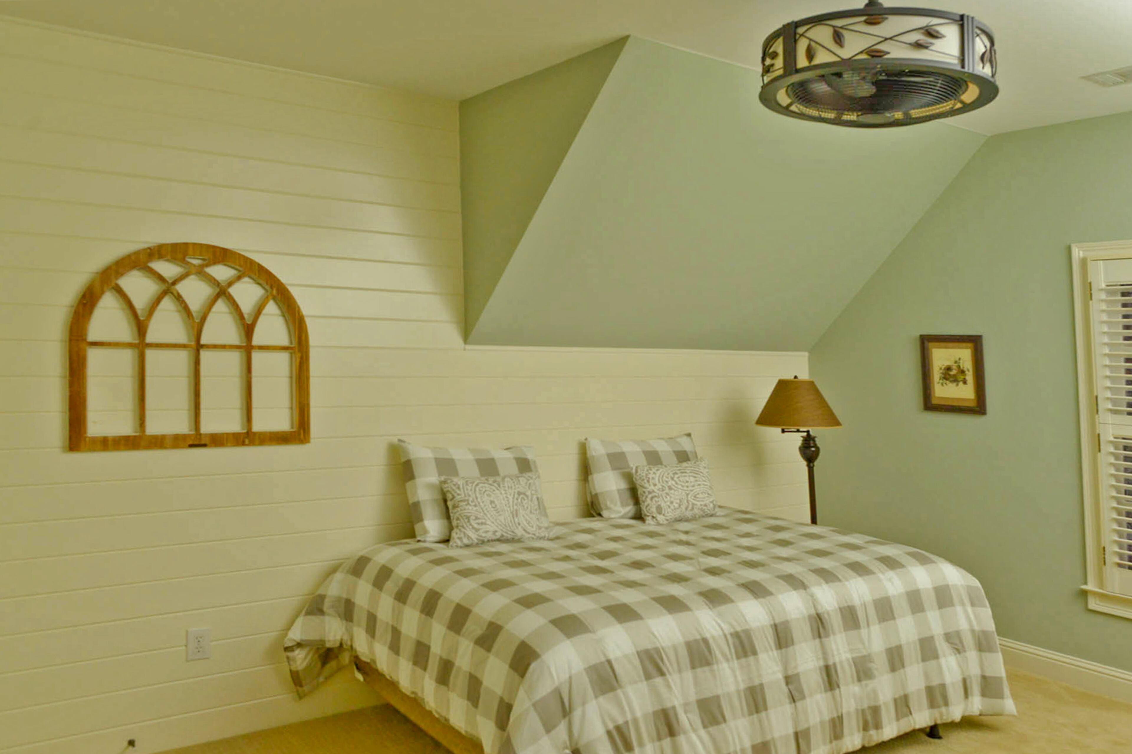 The gingham bedding complements the style of the shiplapped accent wall, which was added in the renovations. A metal dome fan speckled in leaves brings a sense of the outdoors within the space while the art piece is reminiscent of a trellis.