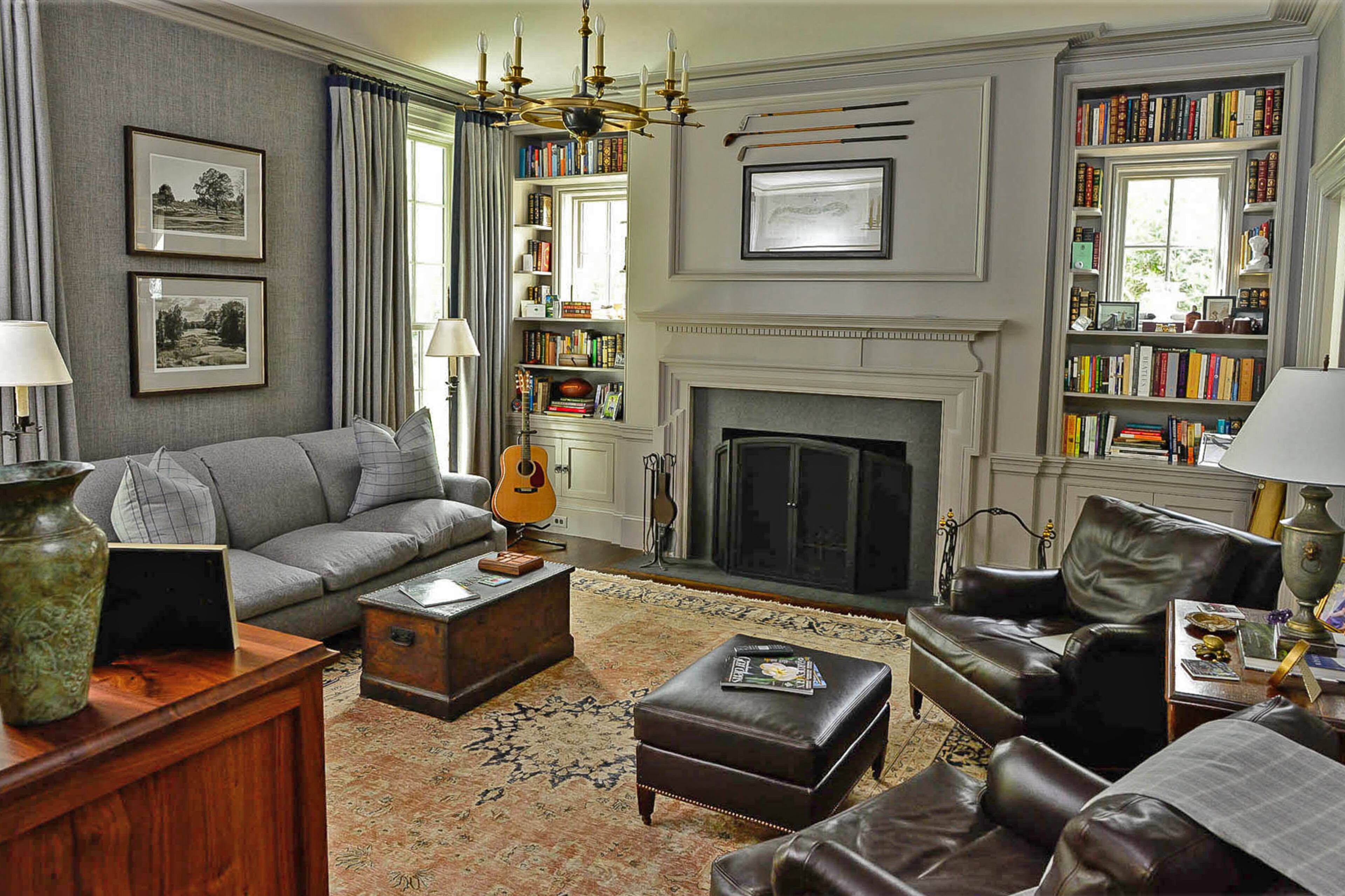 The study is awash in gray, from the grasscloth wallpaper to the painted trim and millwork and the upholstery. The bookshelves by Kyle Priest with Rosewood Millwork were designed around windows to bring in natural light. Above the fireplace, wooden shaft golf clubs join with artwork from St. Andrews Links in Scotland.
