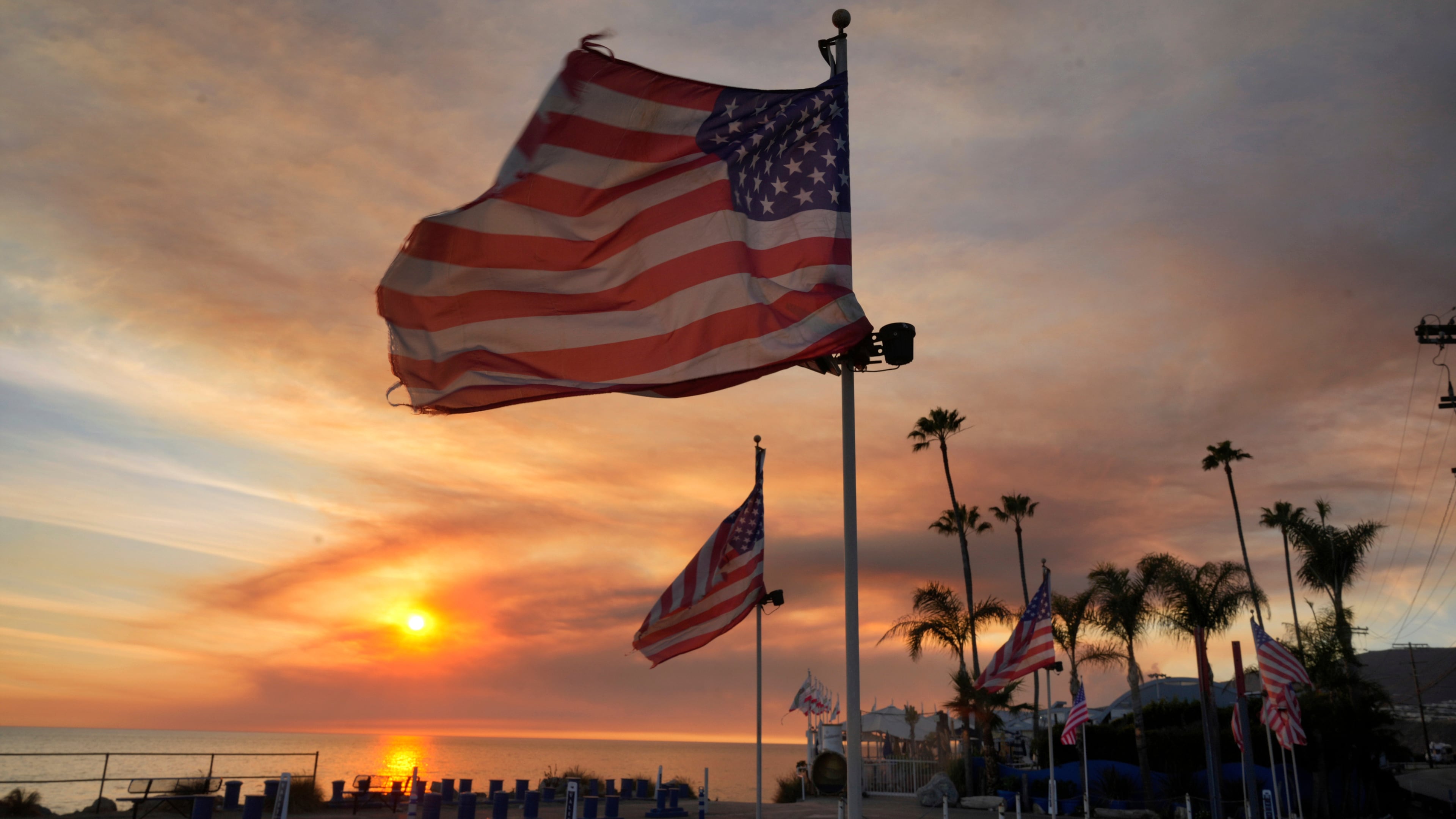 FILE - Flags fly under heavy winds before sunset as a plume of smoke from the Franklin Fire rises over the ocean Tuesday, Dec. 10, 2024, in Malibu, Calif. (AP Photo/Damian Dovarganes,File)