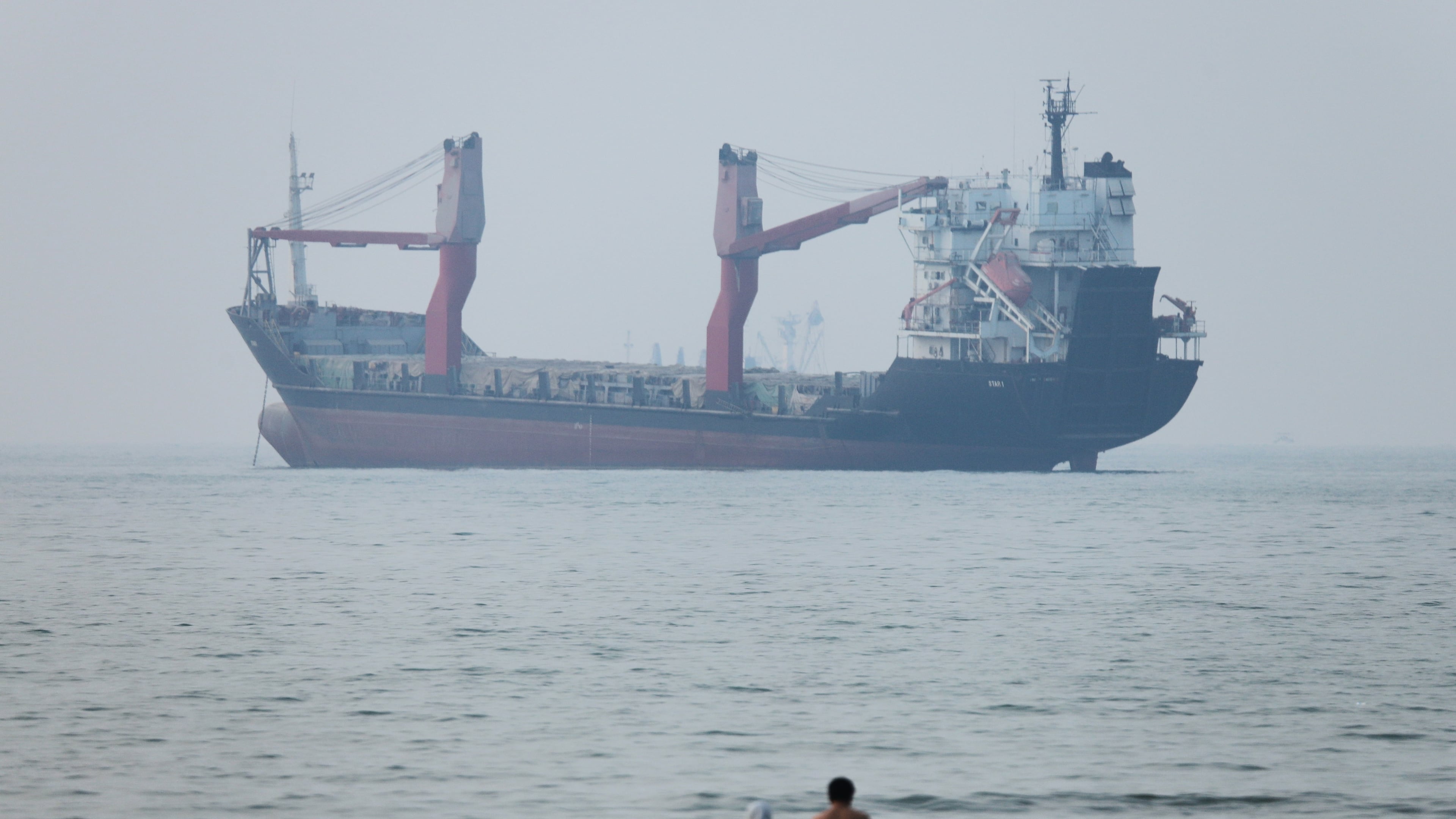 A group of people stands in shallow water as a cargo ship appears anchored in the Strait of Hormuz off Bandar Abbas, Iran, Tuesday, June 30, 2026. (Amirhosein Khorgooi/ISNA via AP)