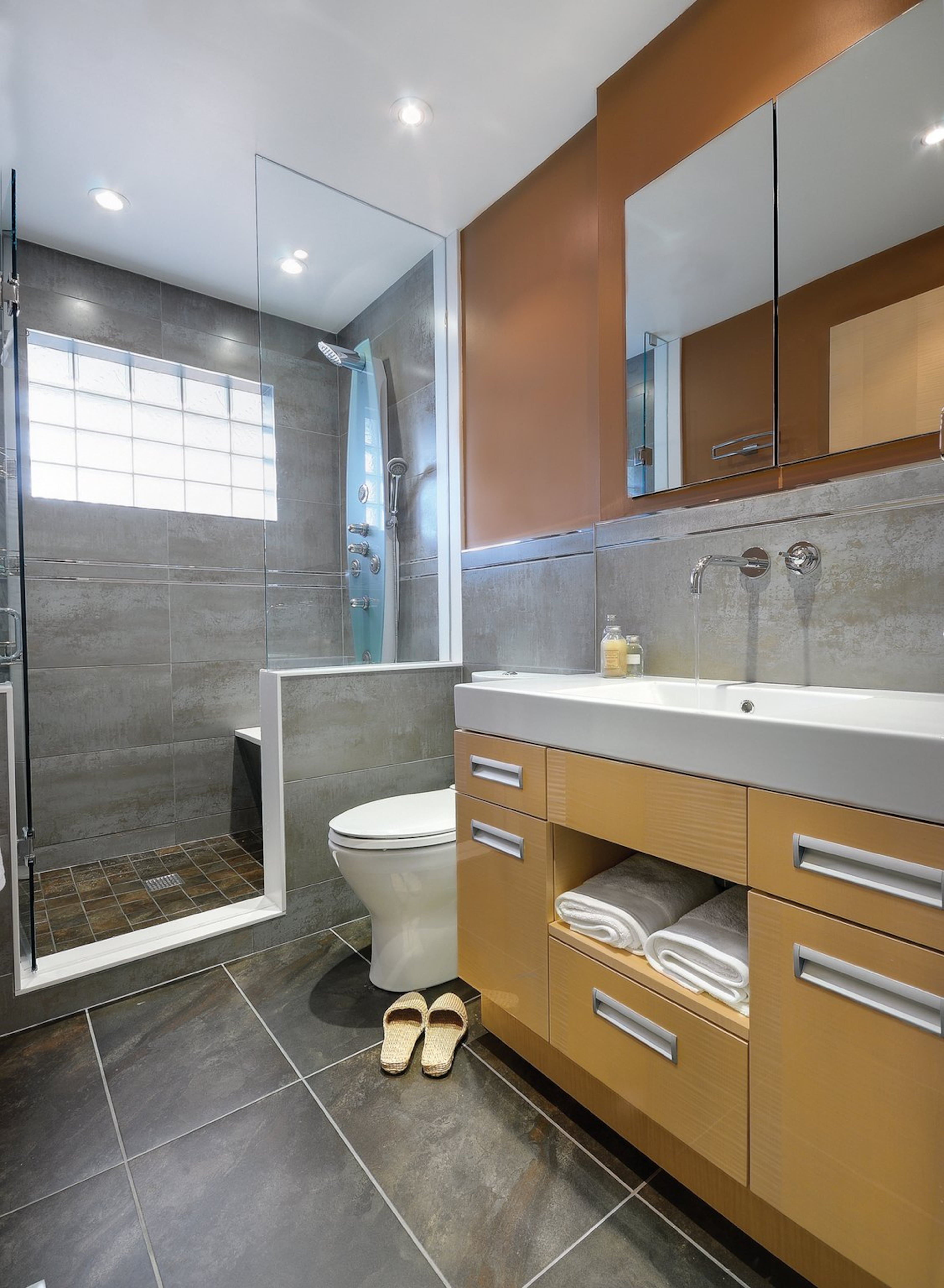 A generous shower with unique water features, an ice glass block window and a convenient seat replaced a traditional tub. Natural sycamore cabinetry, in a high-gloss finish, offers a warm contrast to the floor and wall tiles.